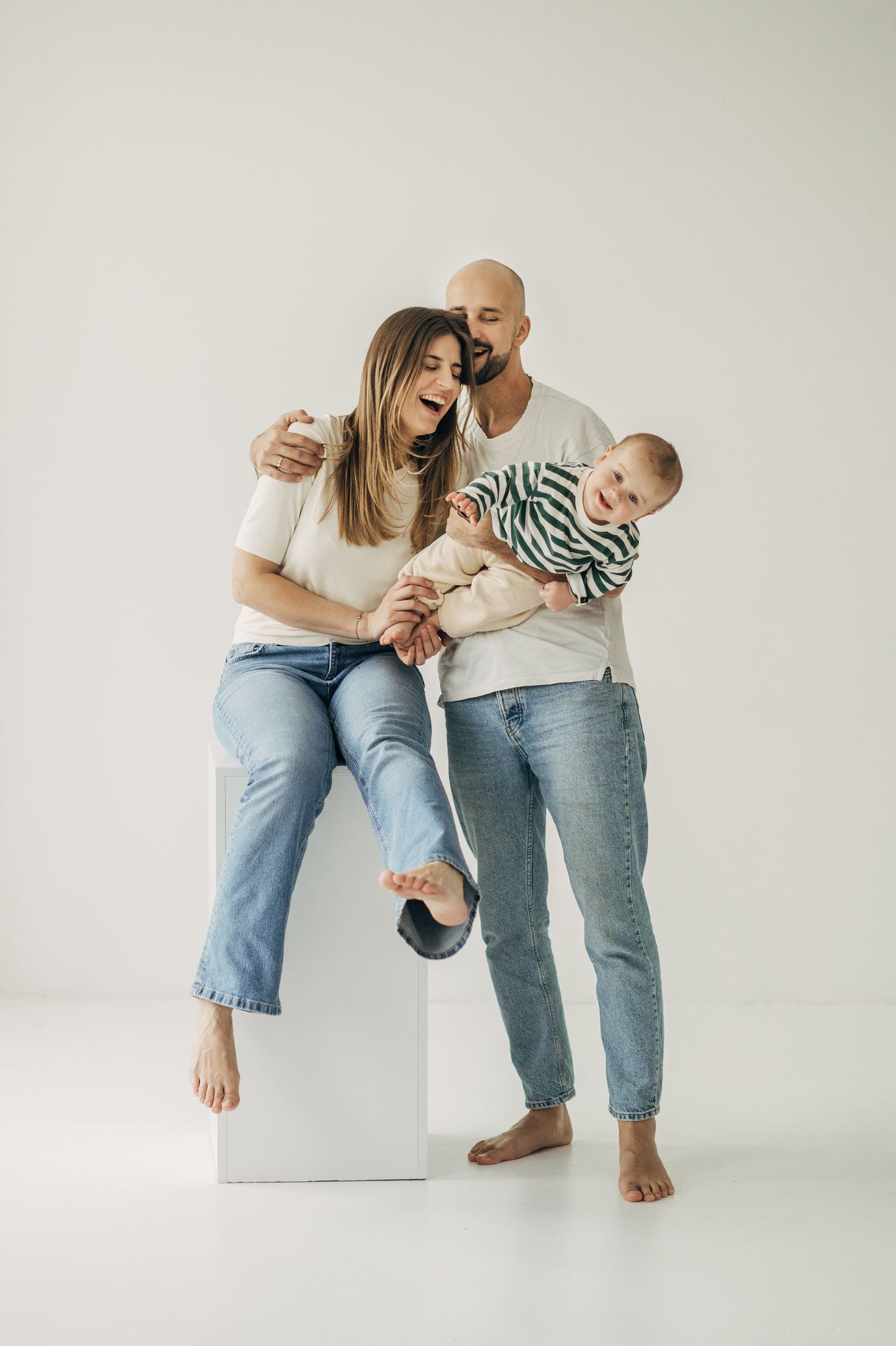 Family in casual denim outfits poses playfully on white cube seats against light background.