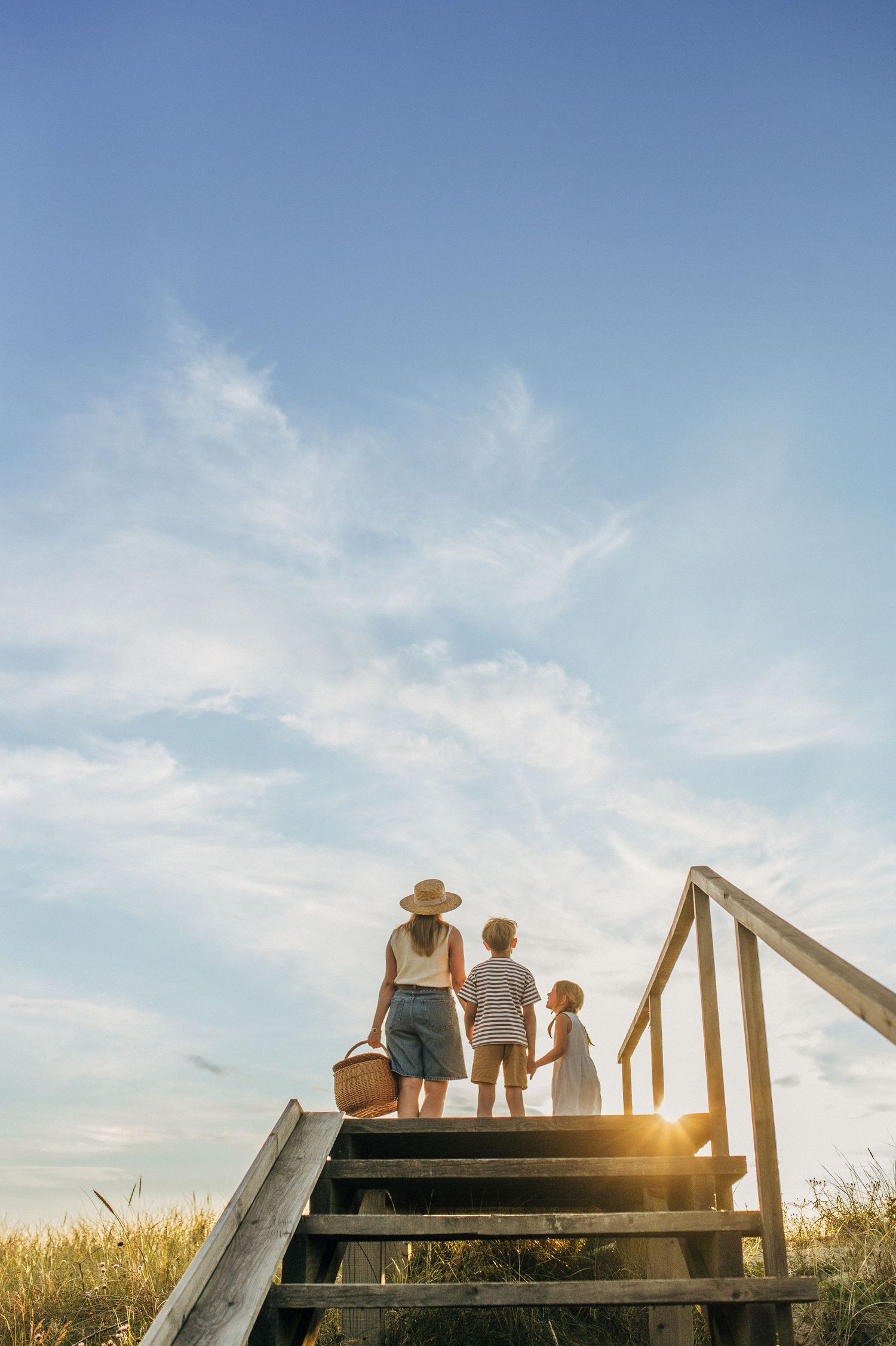 A mother and two children climb wooden beach stairs at golden hour, carrying a picnic basket under a blue sky.