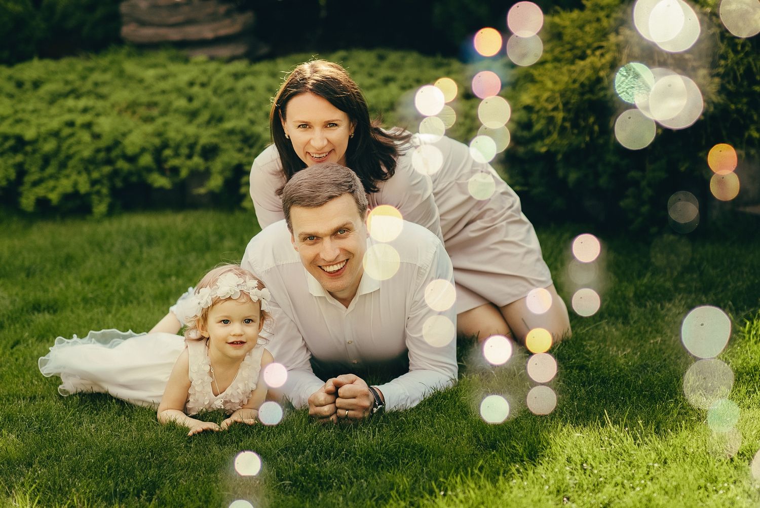 Happy family of three lying on green grass in a garden, parents smiling with young daughter in white dress.