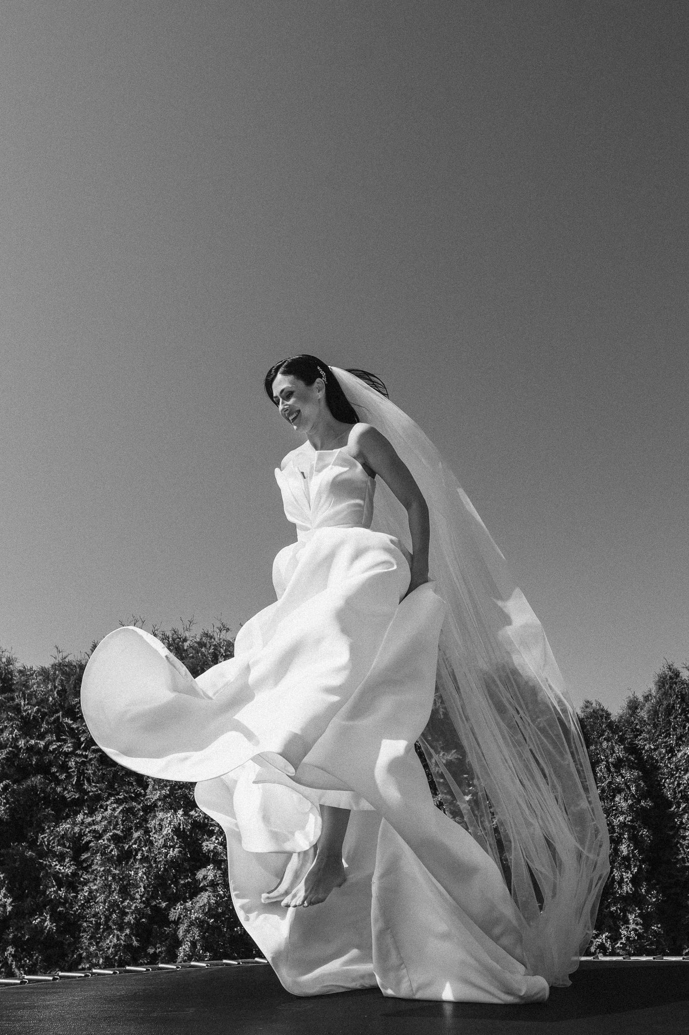 A bride in a flowing white gown and veil twirls joyfully outdoors in a black and white photograph.