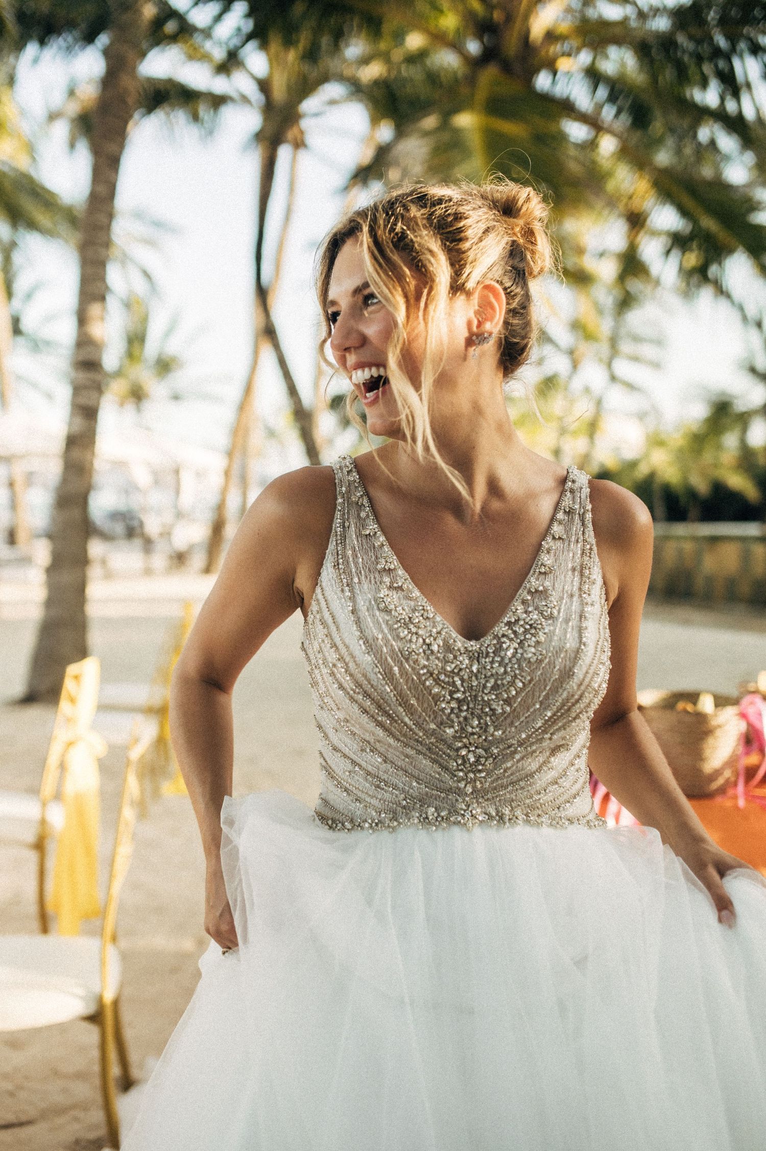 A wedding dress with a beaded v-neck bodice and flowing tulle skirt sparkles in golden sunlight among palm trees.