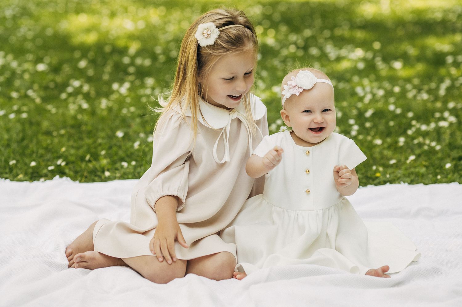 Two little girls in white and beige dresses sit together on blanket in flower-filled meadow.