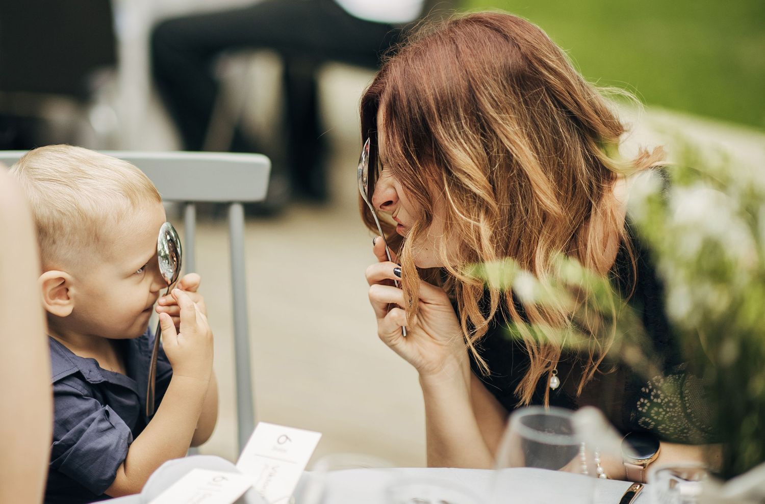 Young mother bonding with toddler at outdoor cafe, child holding spoon while mom smiles closely.