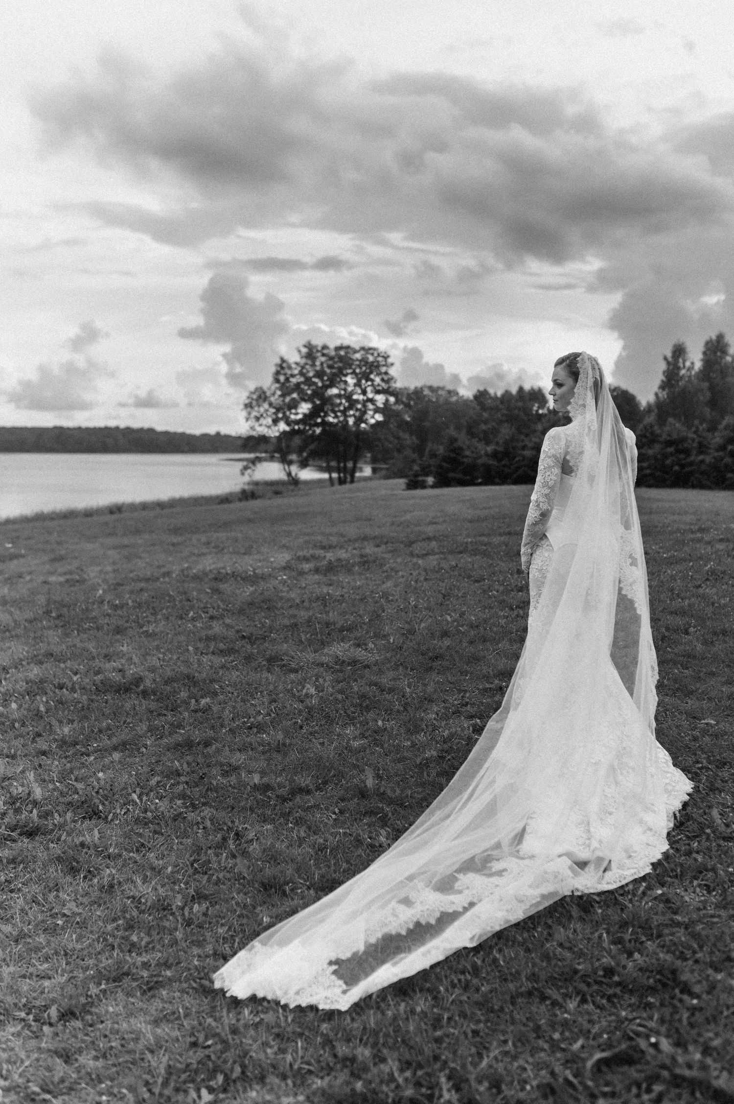 A flowing white wedding veil trails dramatically across a grassy hilltop field under moody clouds in black and white.