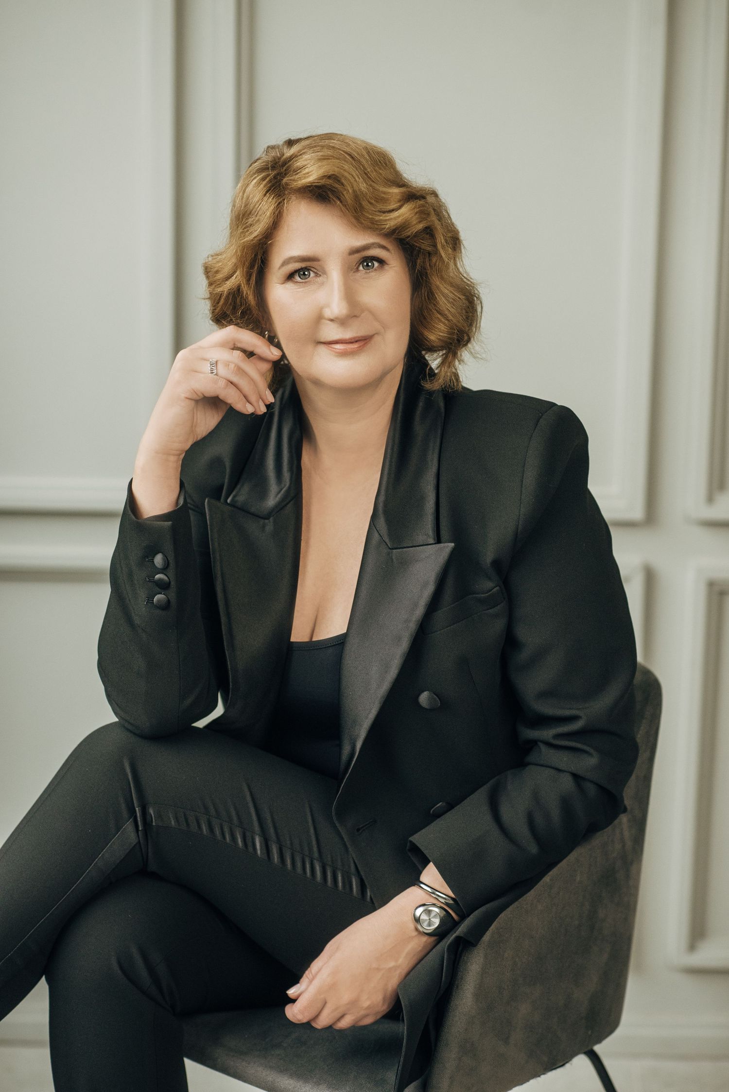 Professional portrait in black business suit seated in dark chair against light background.