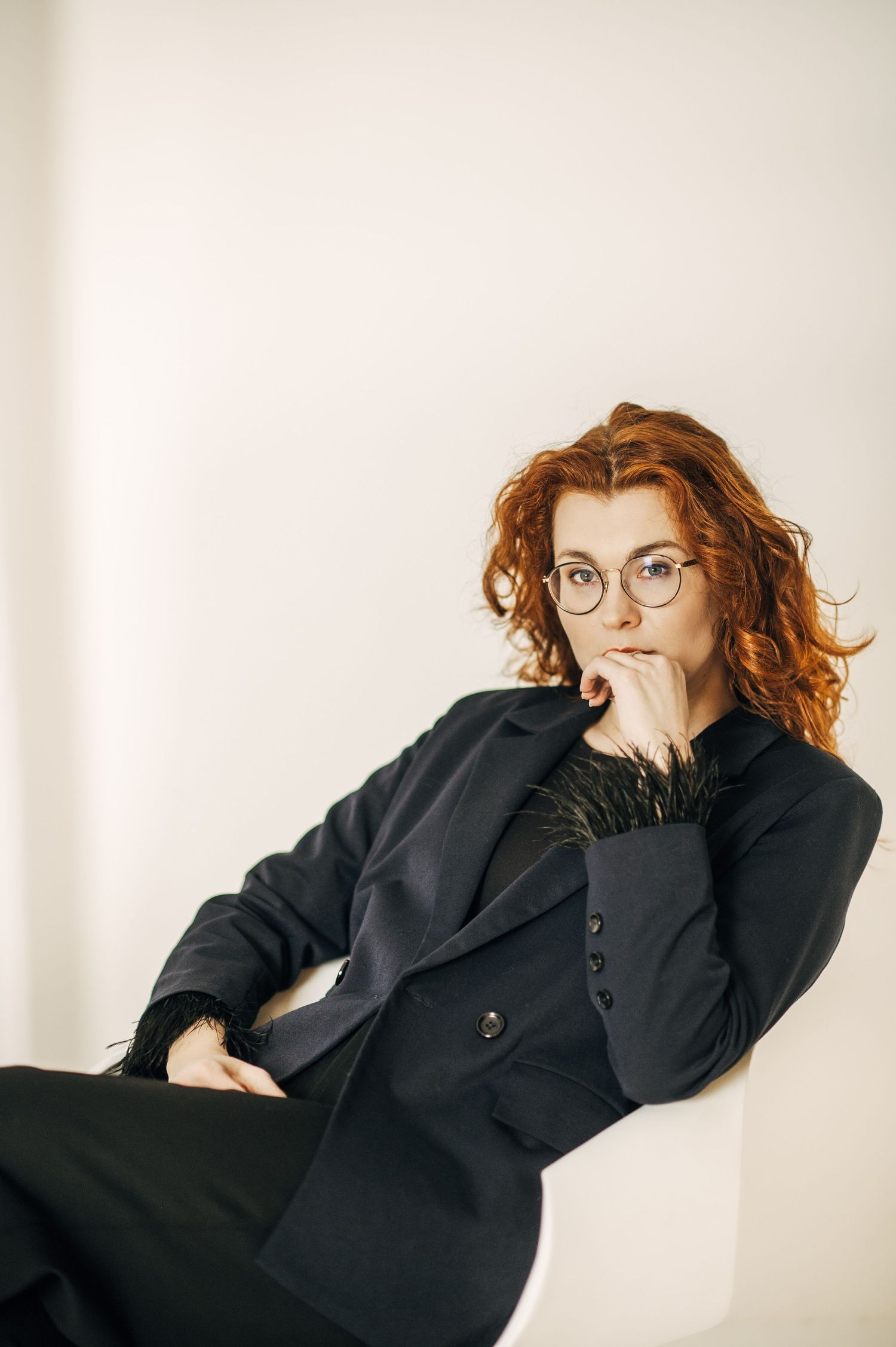 Professional portrait of person in black blazer and glasses with curly red hair against white background.