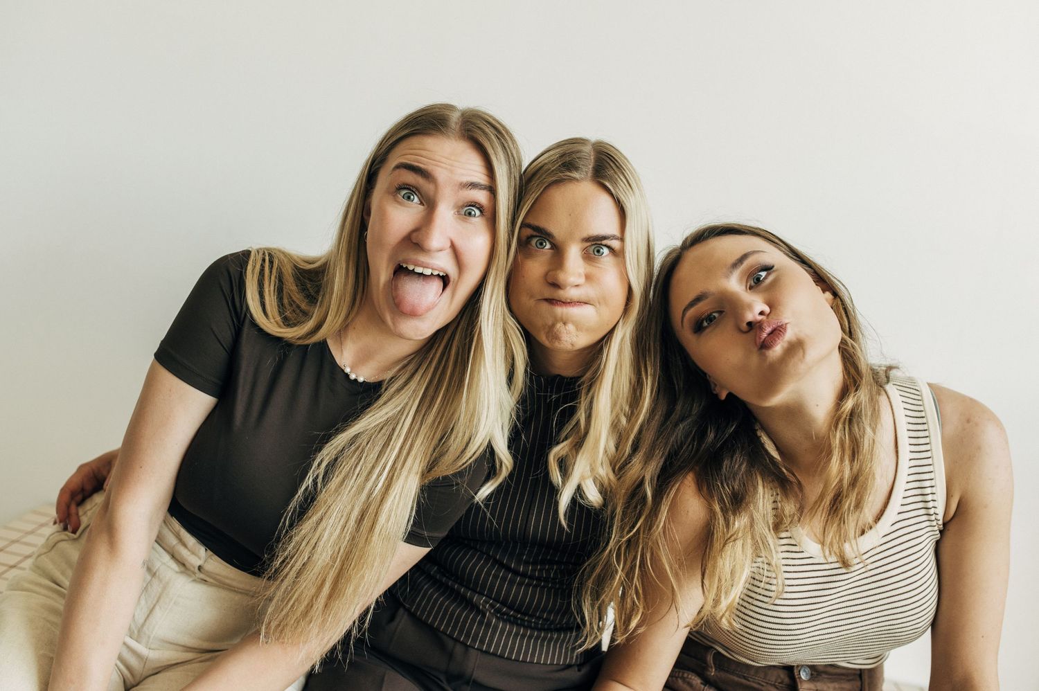 Three young blonde women making silly faces together, sticking out tongues and pouting, sitting against a white wall.