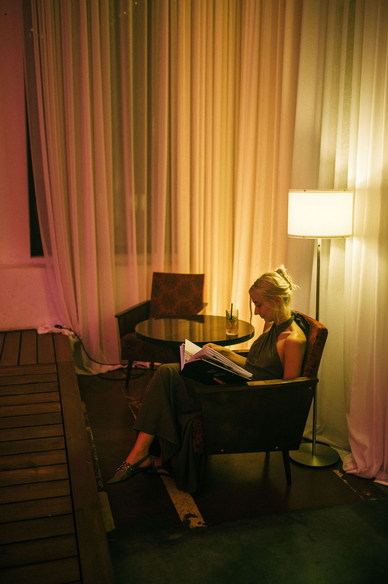 Woman reading a magazine in a cozy armchair beside a glowing floor lamp with warm curtains backdrop.