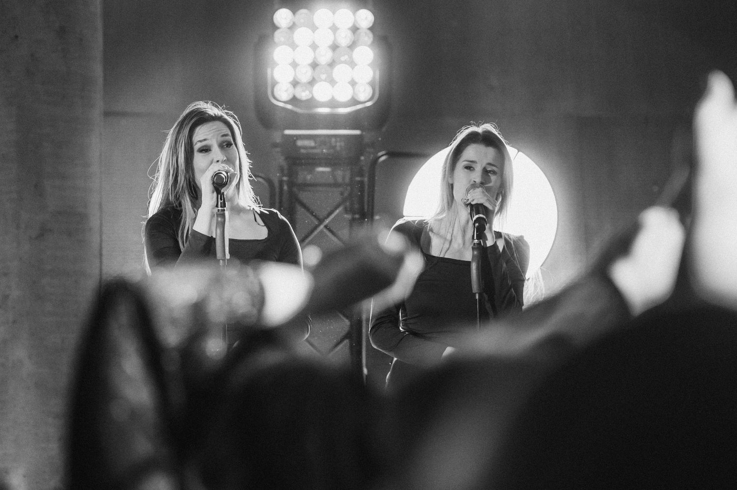 Black and white concert photo showing performers singing into microphones with dramatic spotlights behind them on stage.