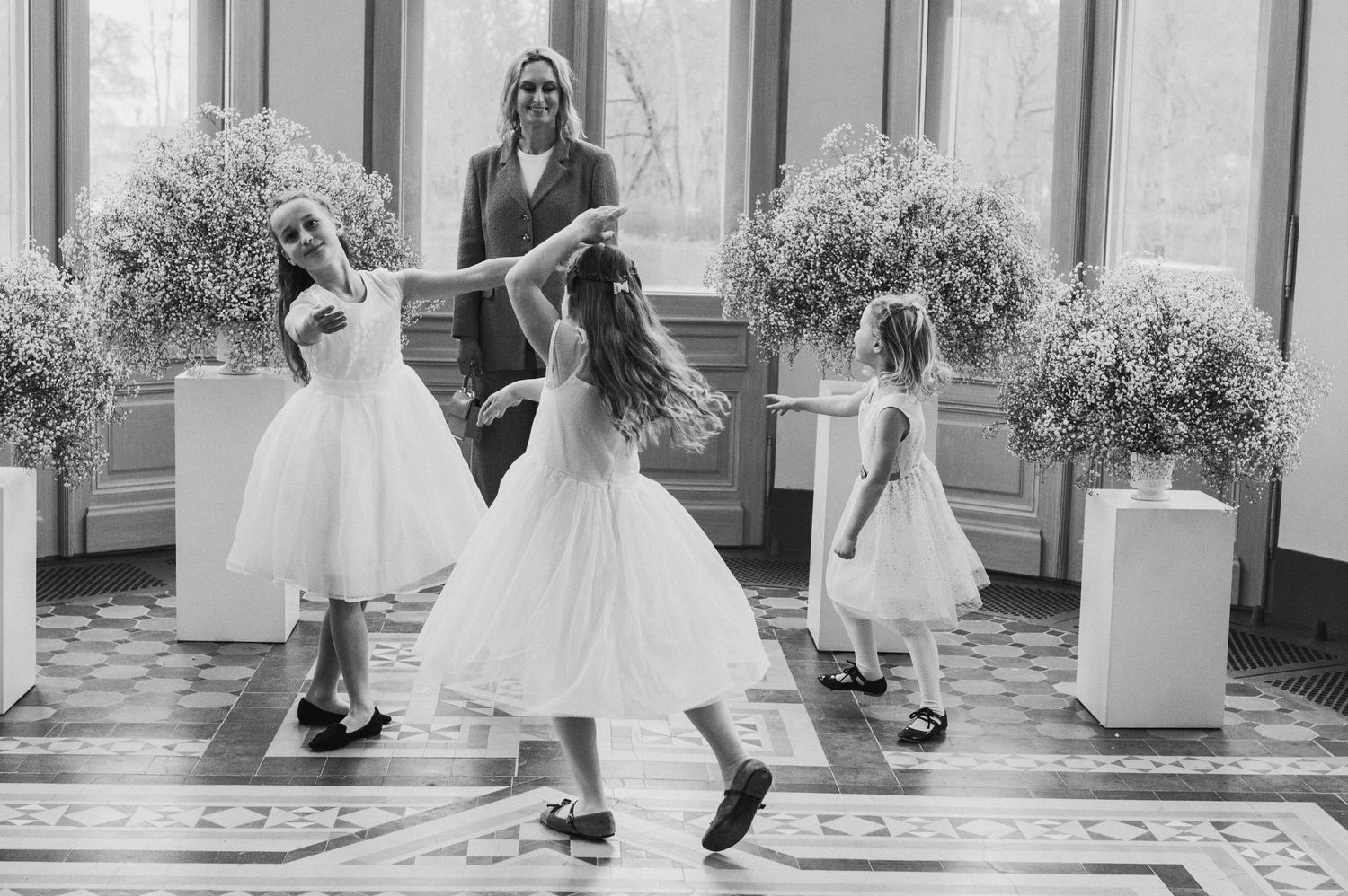 Black and white photo of flower girls in white dresses dancing with a woman in an elegant floral-decorated hall.