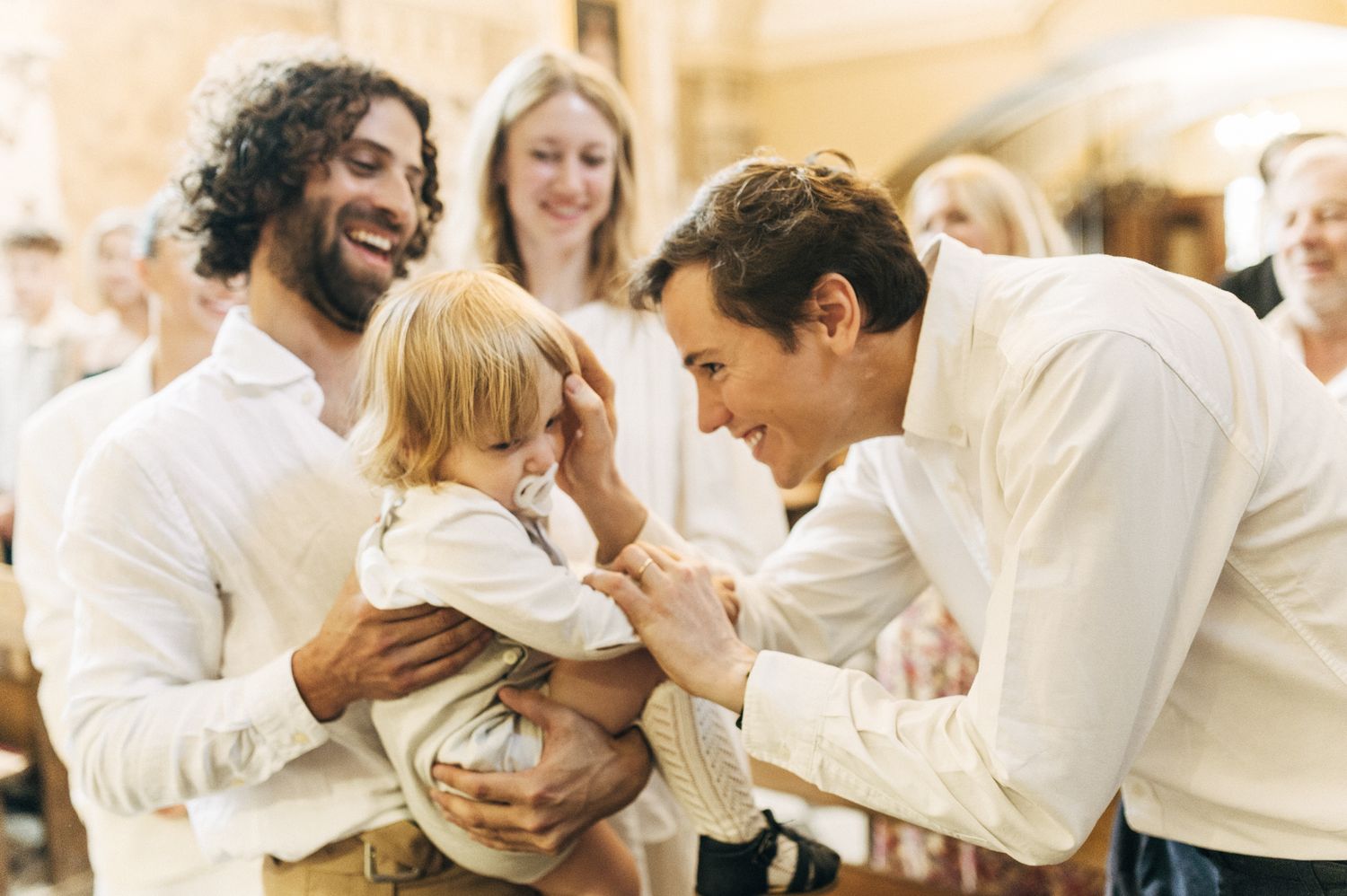 Two smiling men in white shirts interacting warmly with a baby at a christening ceremony.