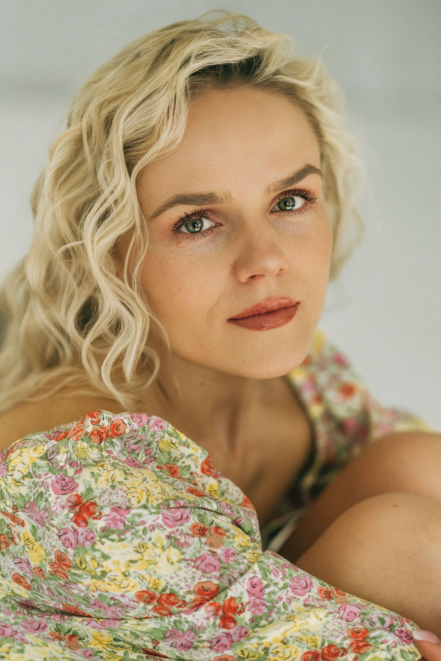 Blonde woman with curly hair wearing a floral dress, gazing thoughtfully at the camera in soft natural light.