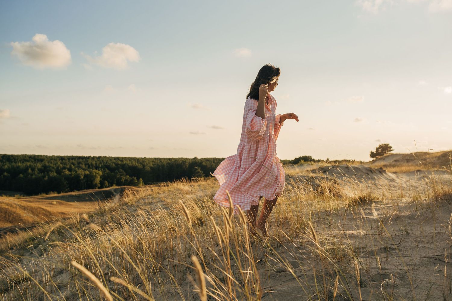 Person in flowing pink dress walking through golden grass at sunset.