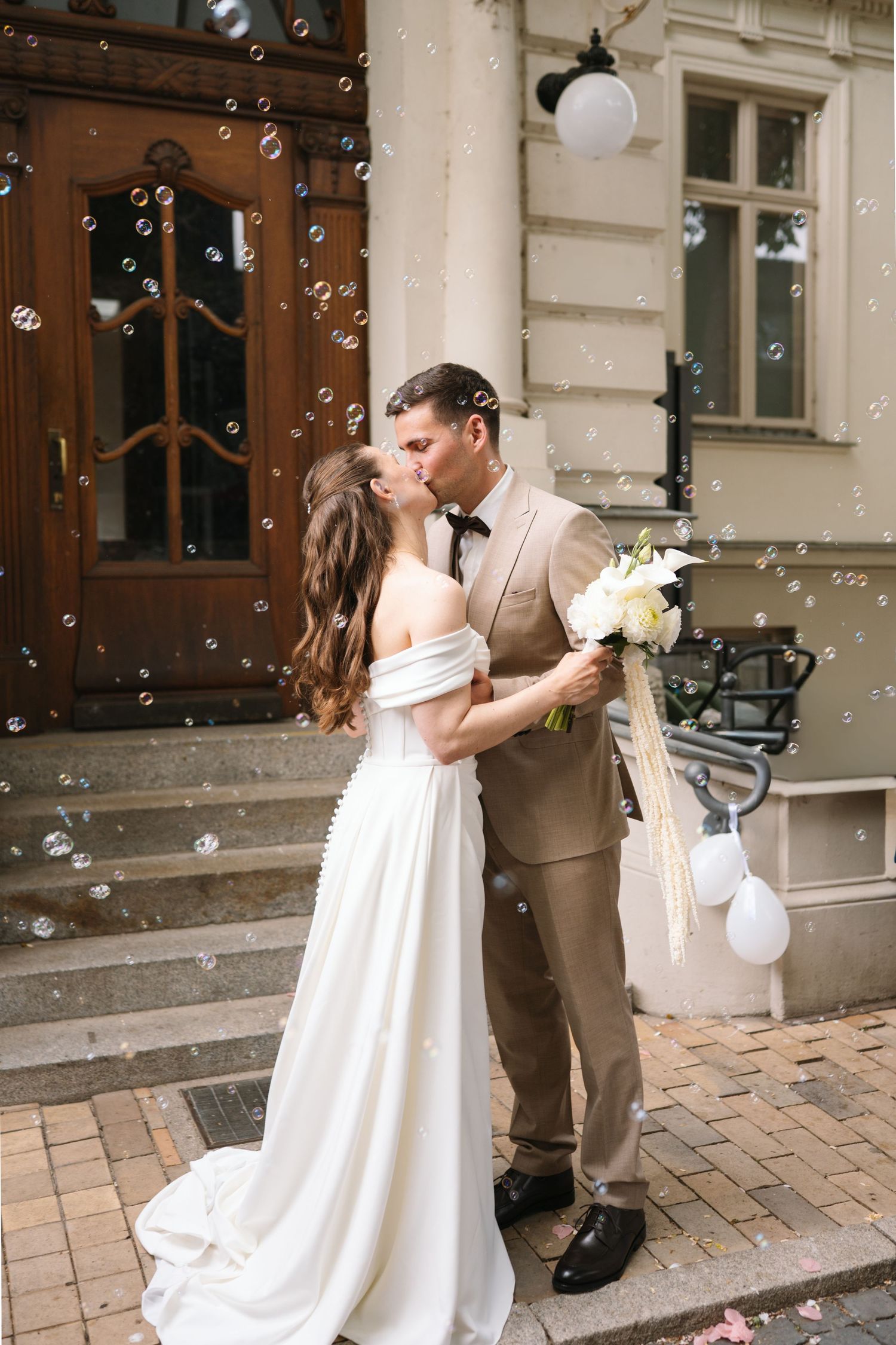 Bride and groom share a romantic kiss on steps surrounded by soap bubbles, bride in white gown holding bouquet.
