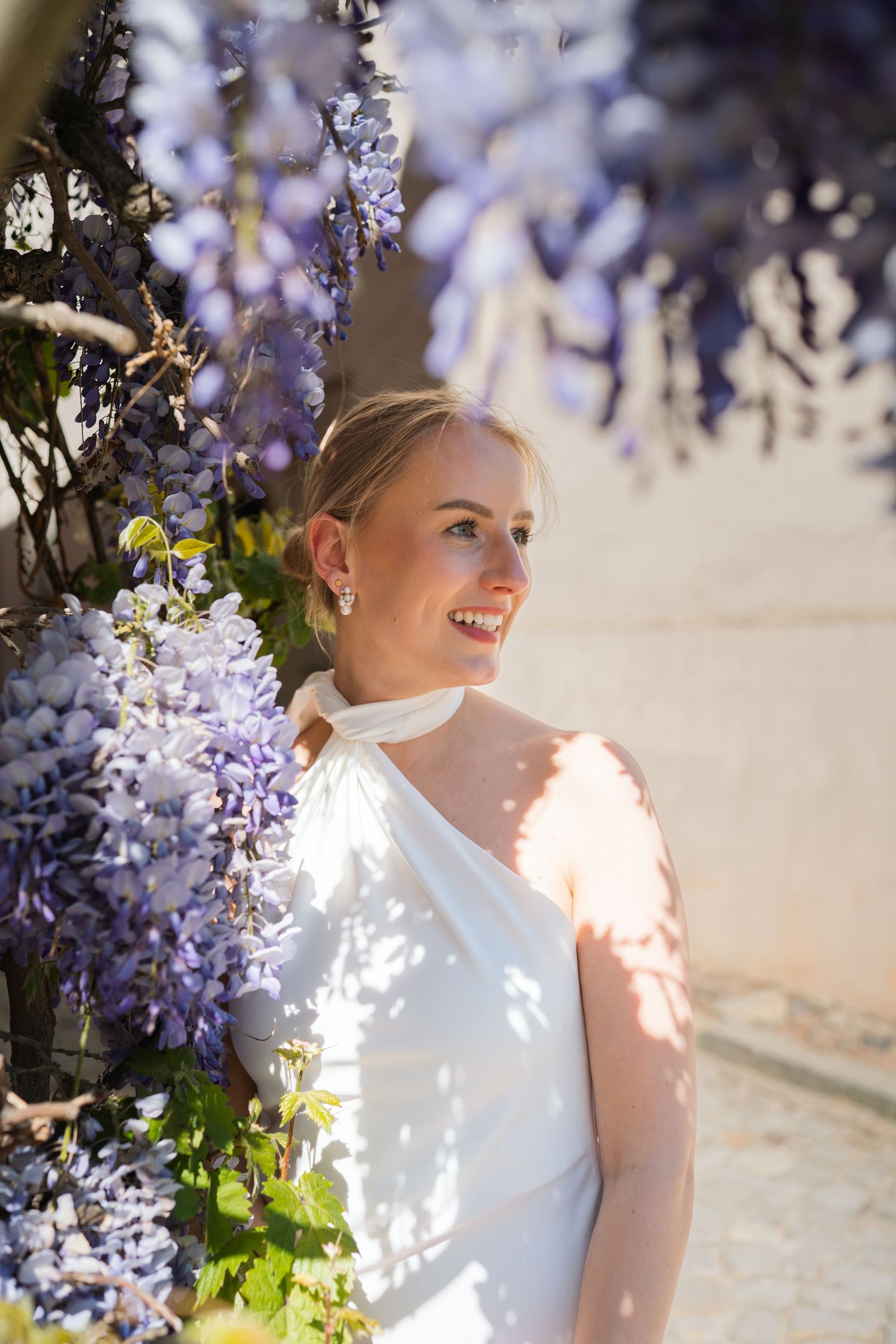 Smiling bride in white dress standing among blooming purple wisteria flowers in warm sunlight.