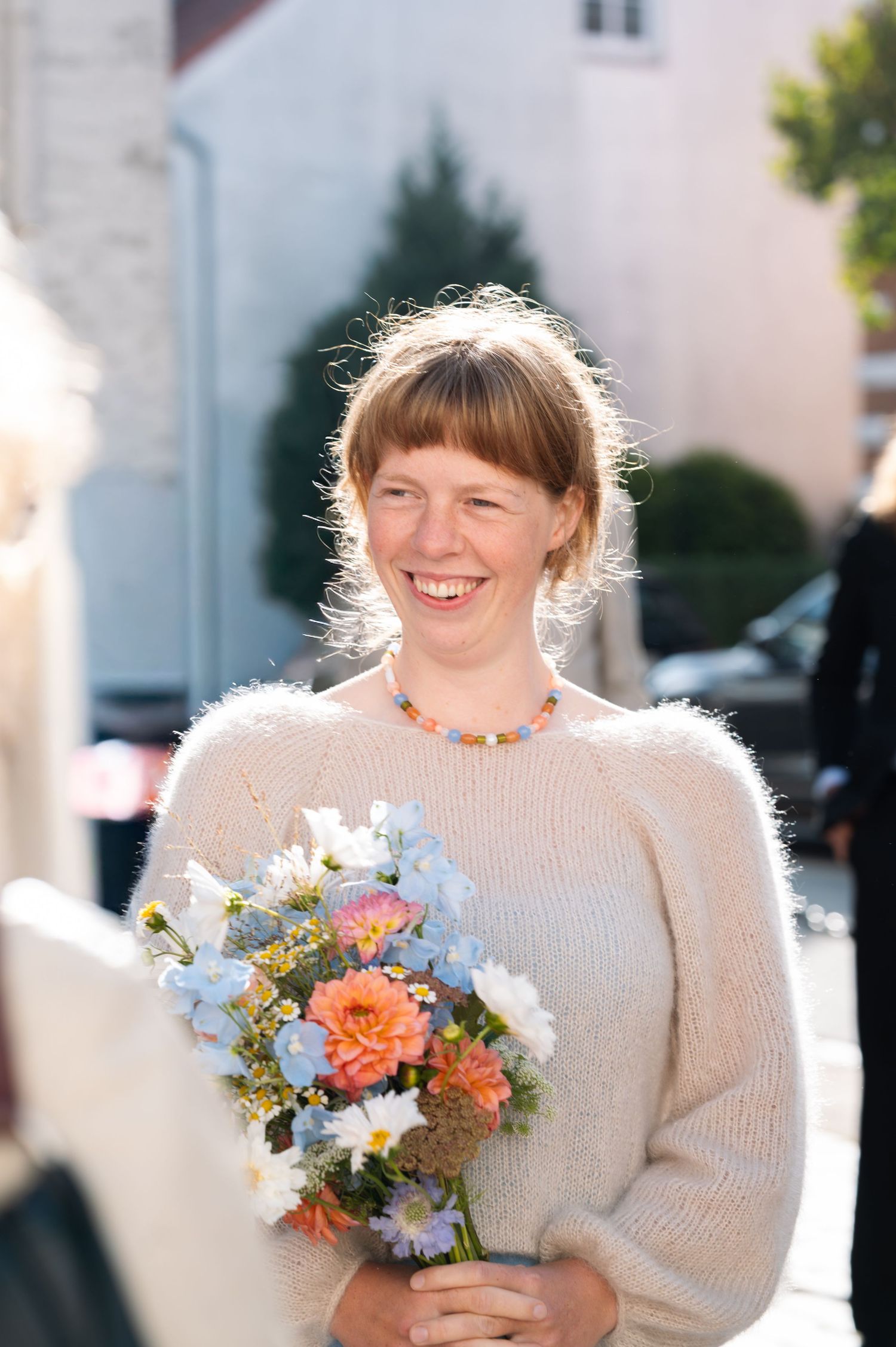 Smiling woman holding colorful flower bouquet outdoors, wearing cream sweater and beaded necklace on sunny day.