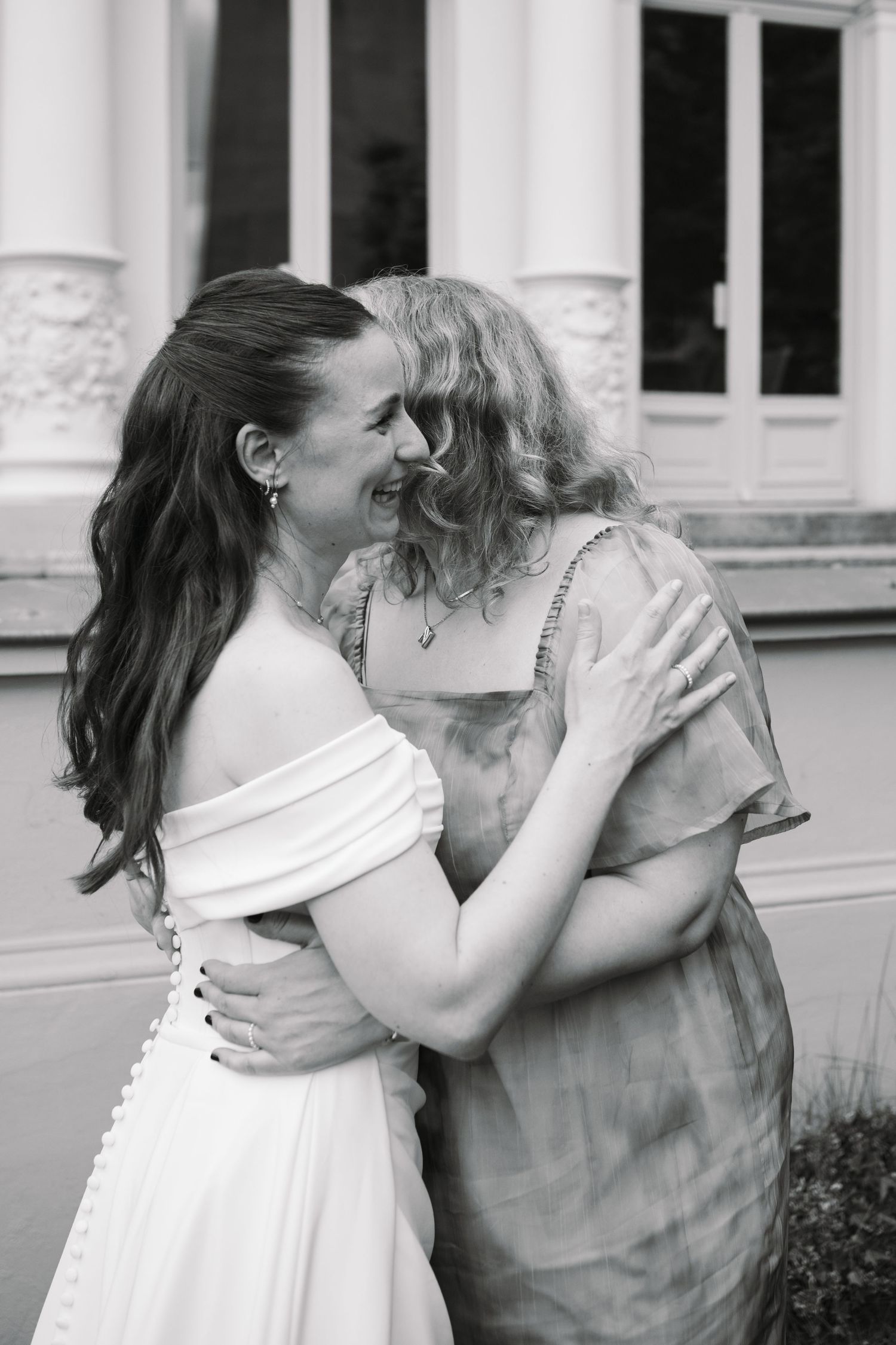 Black and white photo of two women embracing and smiling, one in a white off-shoulder wedding dress.