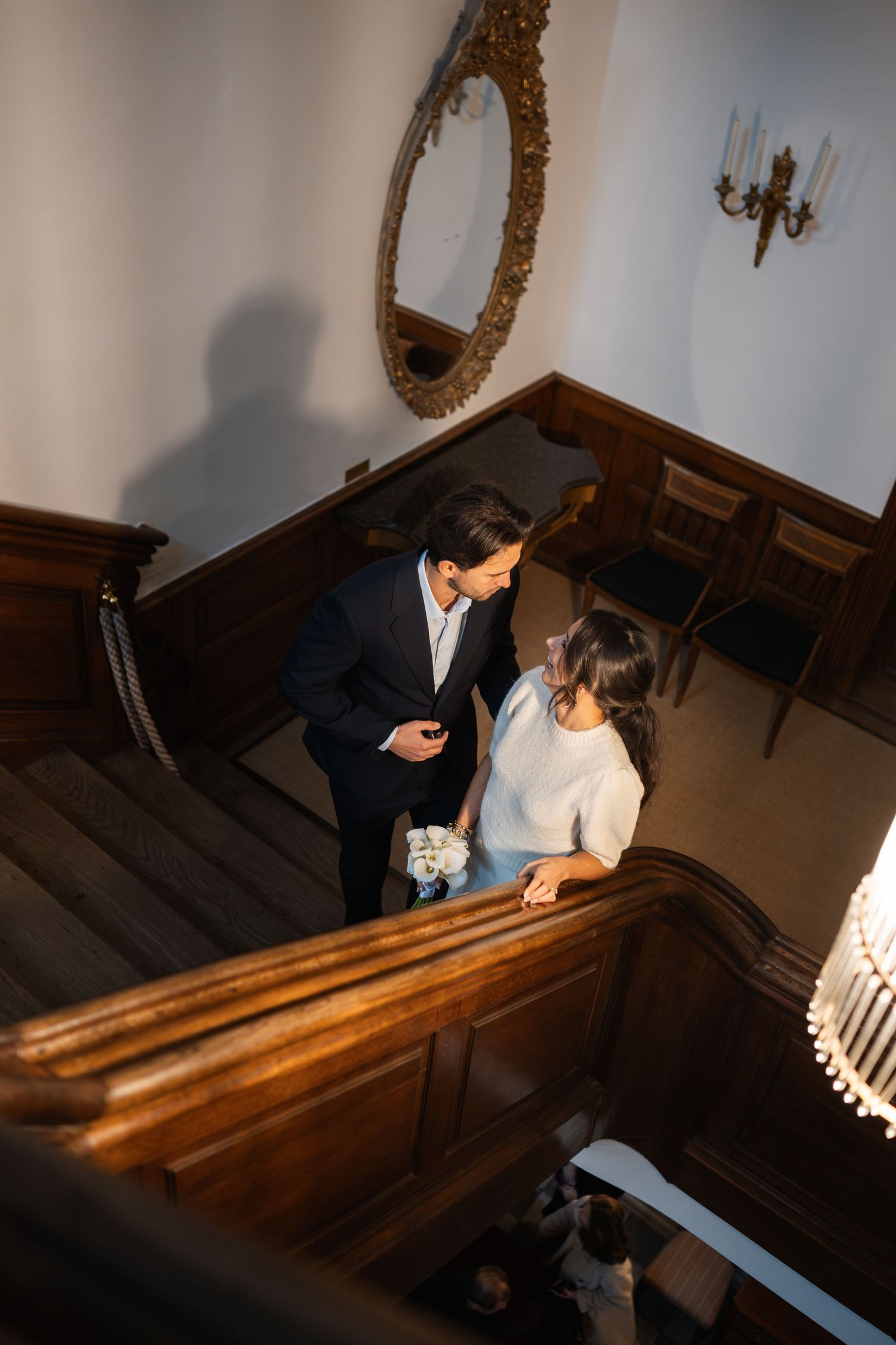 A couple stands together on an elegant wooden staircase with an ornate mirror and chandelier in the background.