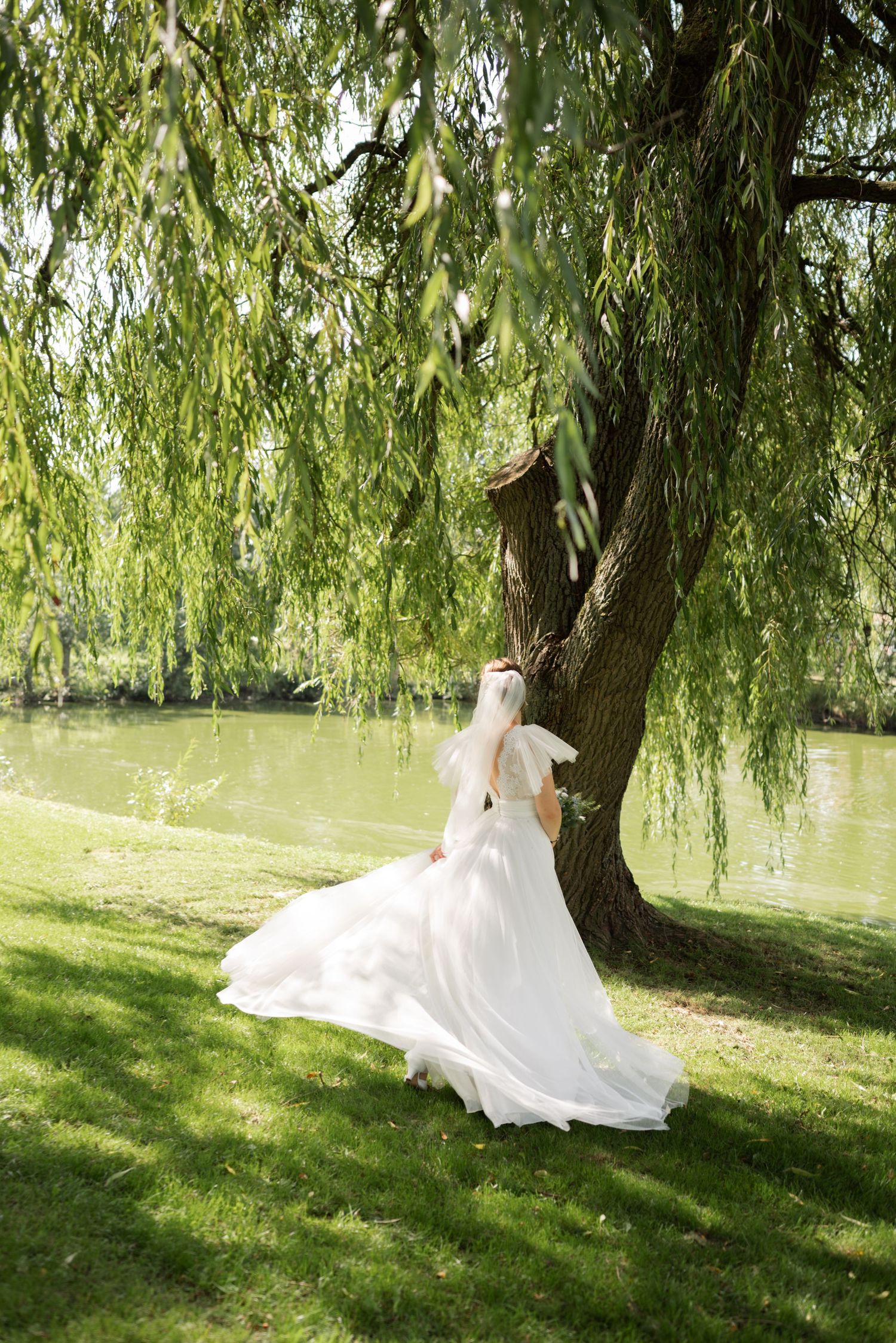 A flowing white wedding dress cascades across green grass beneath a graceful weeping willow tree by a tranquil pond.