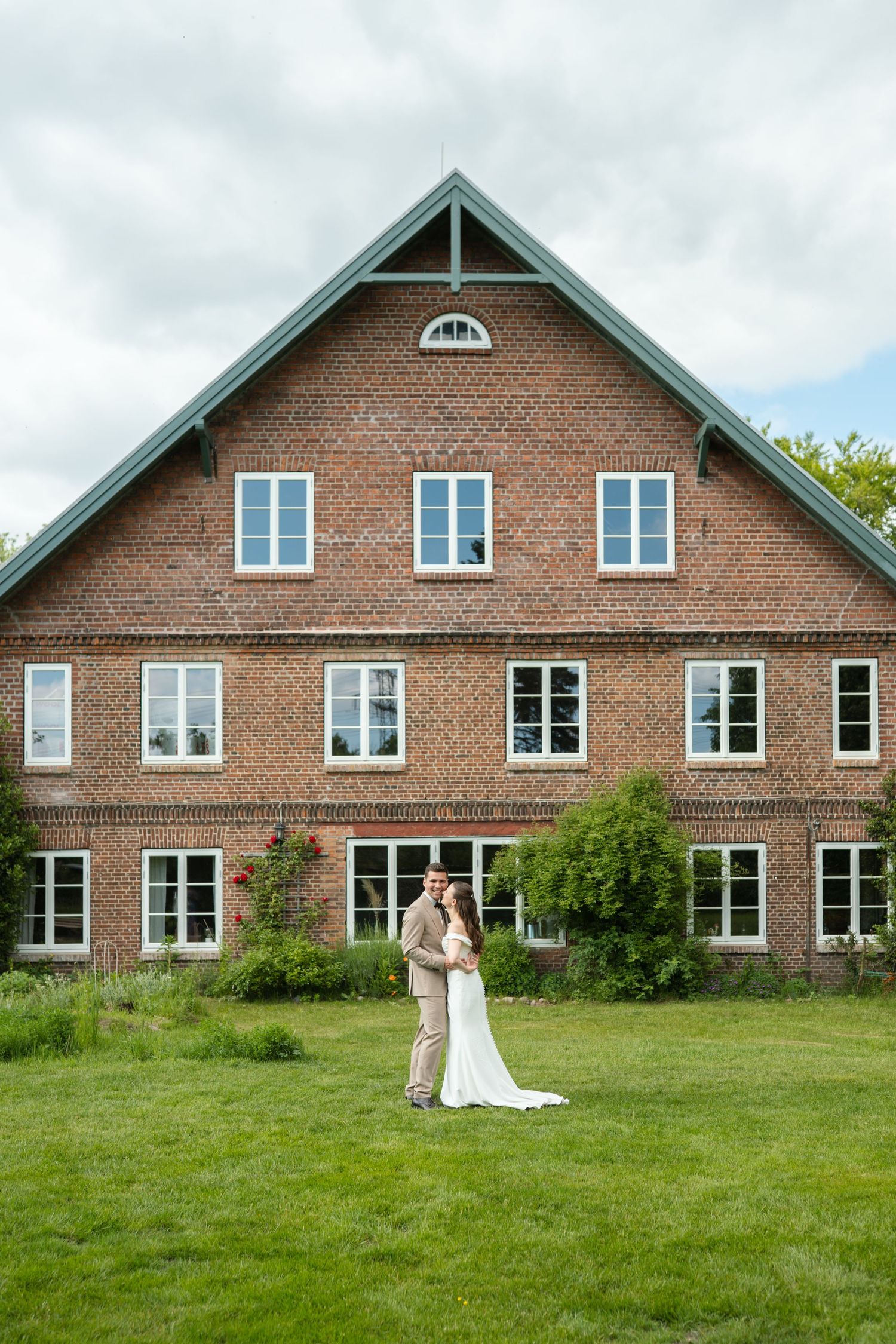 A bride and groom embrace on a green lawn in front of a large red brick country house on a cloudy day.