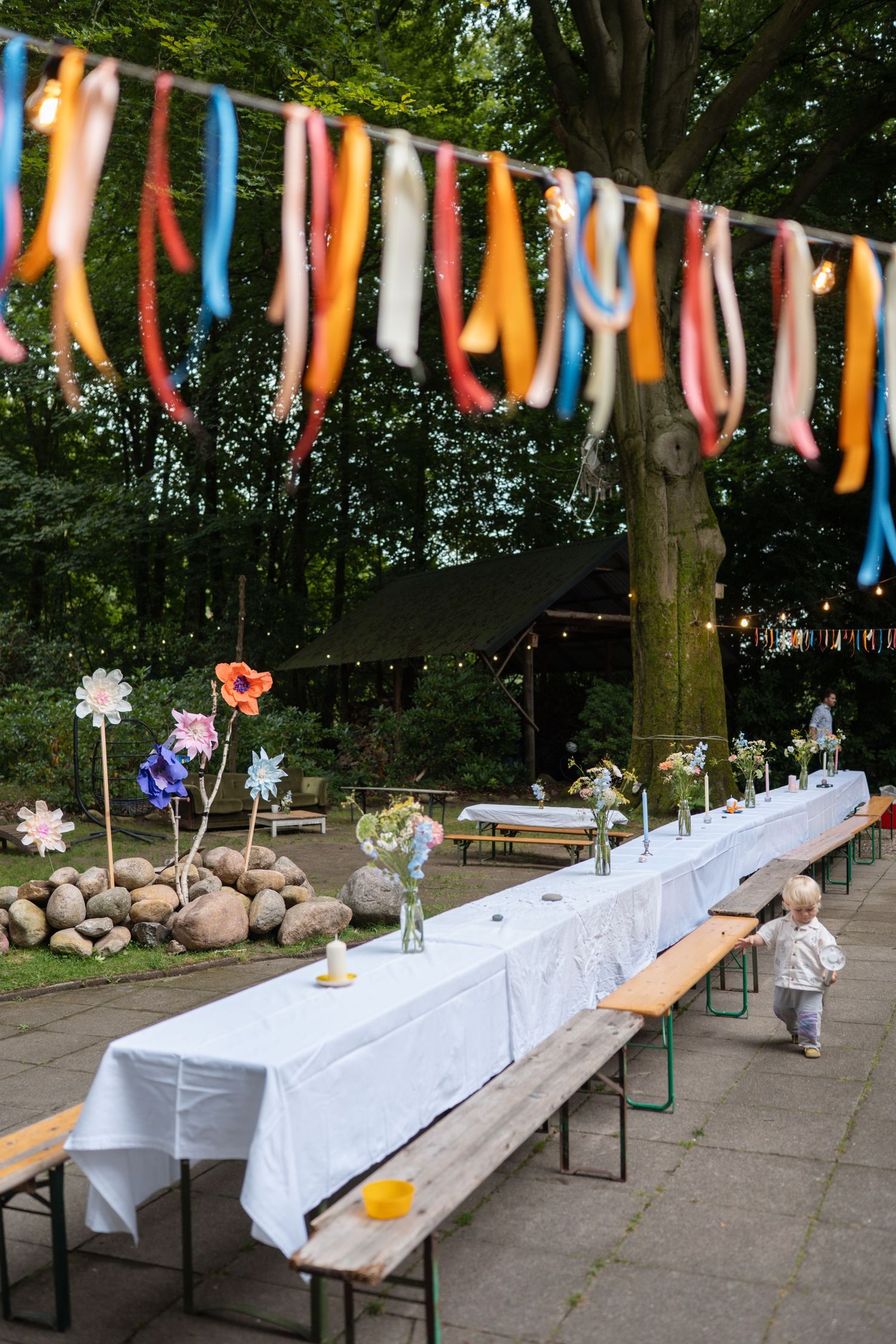 Outdoor party setup with long white tablecloth, wooden benches, colorful ribbon garland, and floral decorations in a garden setting.