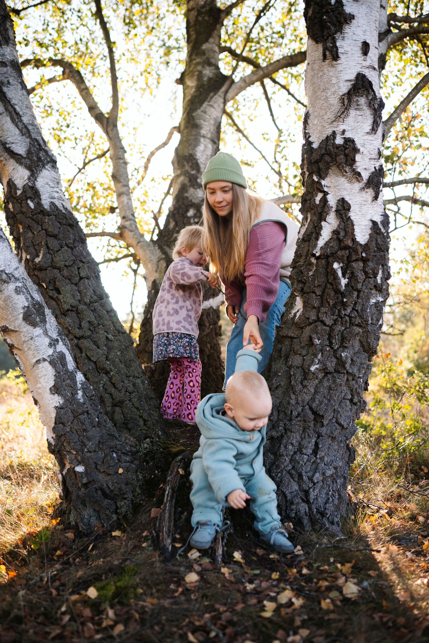 A young woman helps two toddlers climb a birch tree in an autumn forest setting.