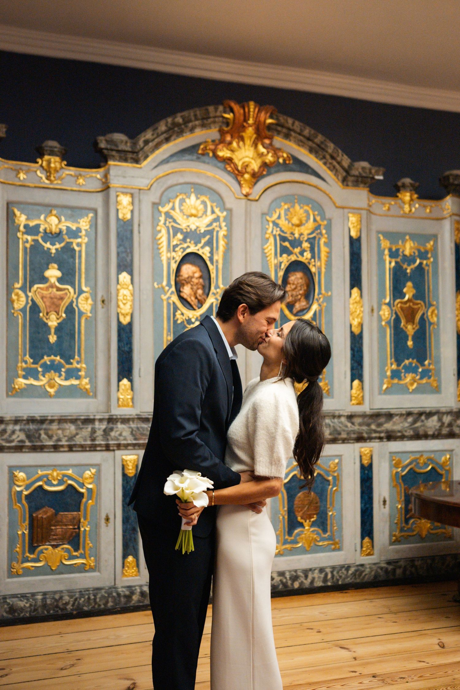 A couple shares a romantic moment in front of an ornate blue and gold baroque wall during their wedding ceremony.