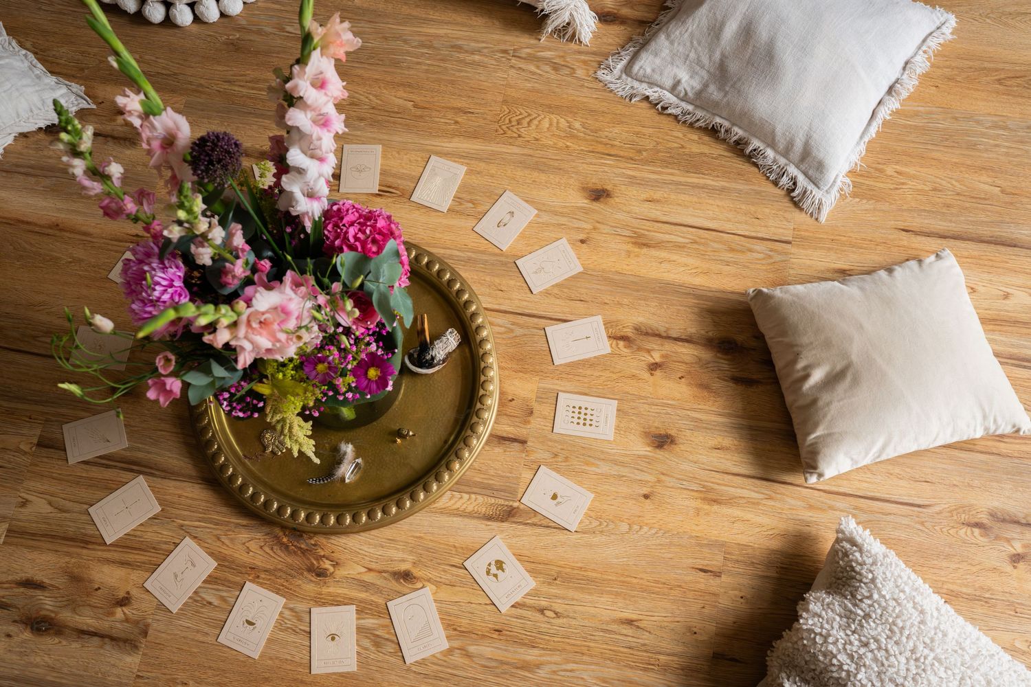 A wooden floor displays scattered tarot cards with decorative cushions and floral arrangements in a mystical setting.