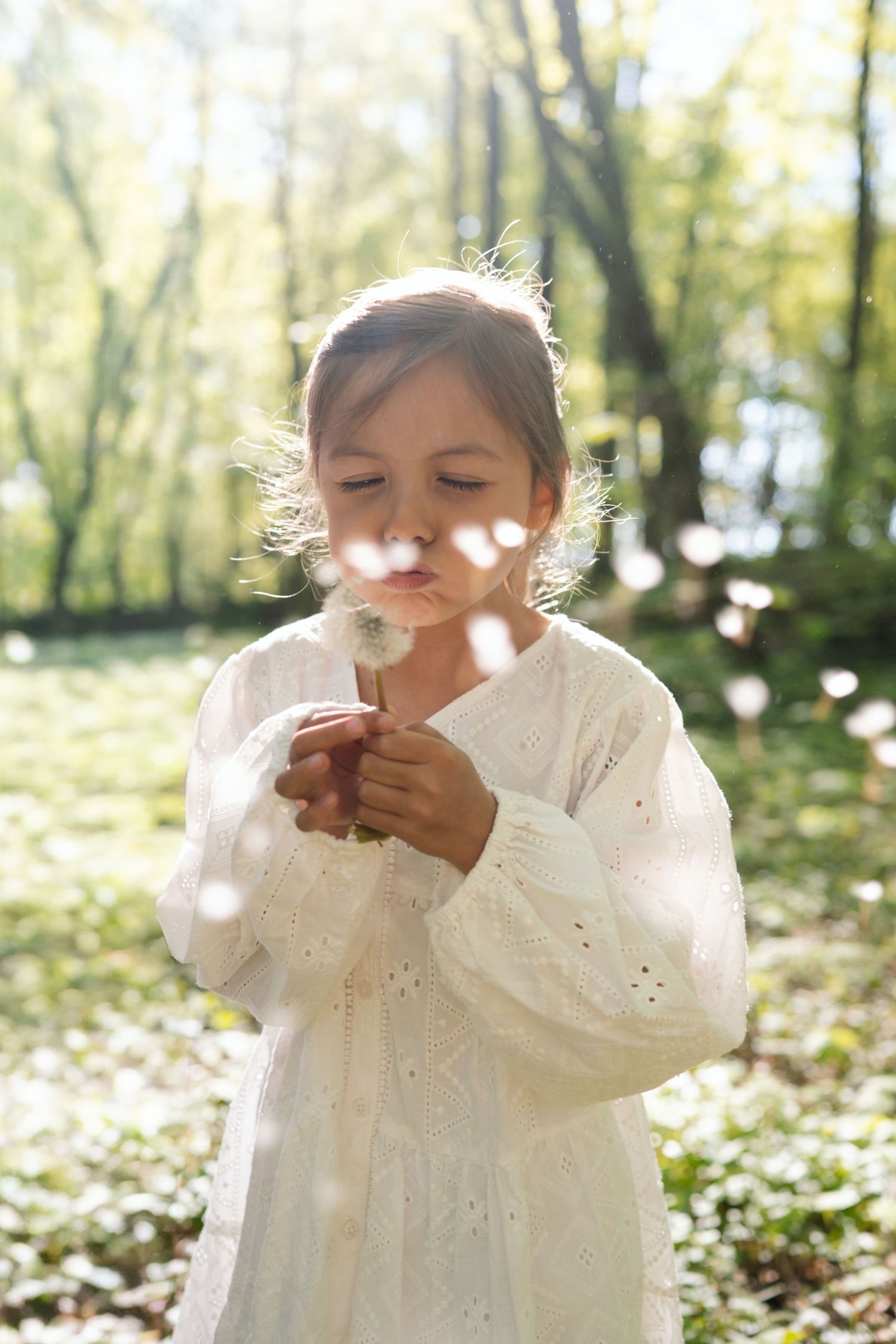 Young girl in white dress blowing dandelion seeds in sunny green forest park.