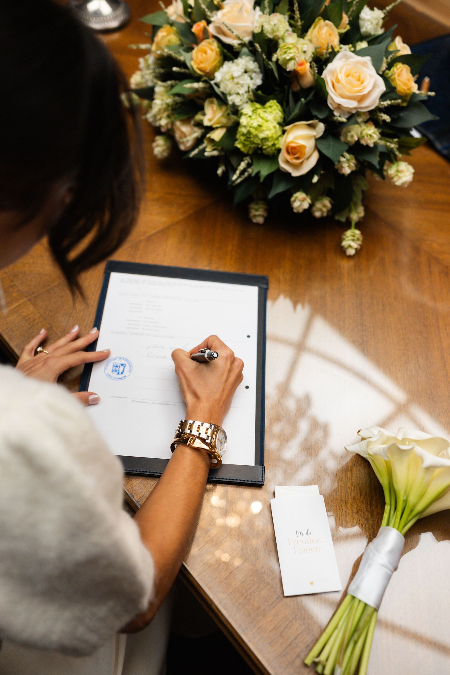 Hands signing wedding documents next to elegant floral bouquets with peach roses on wooden table in natural light.