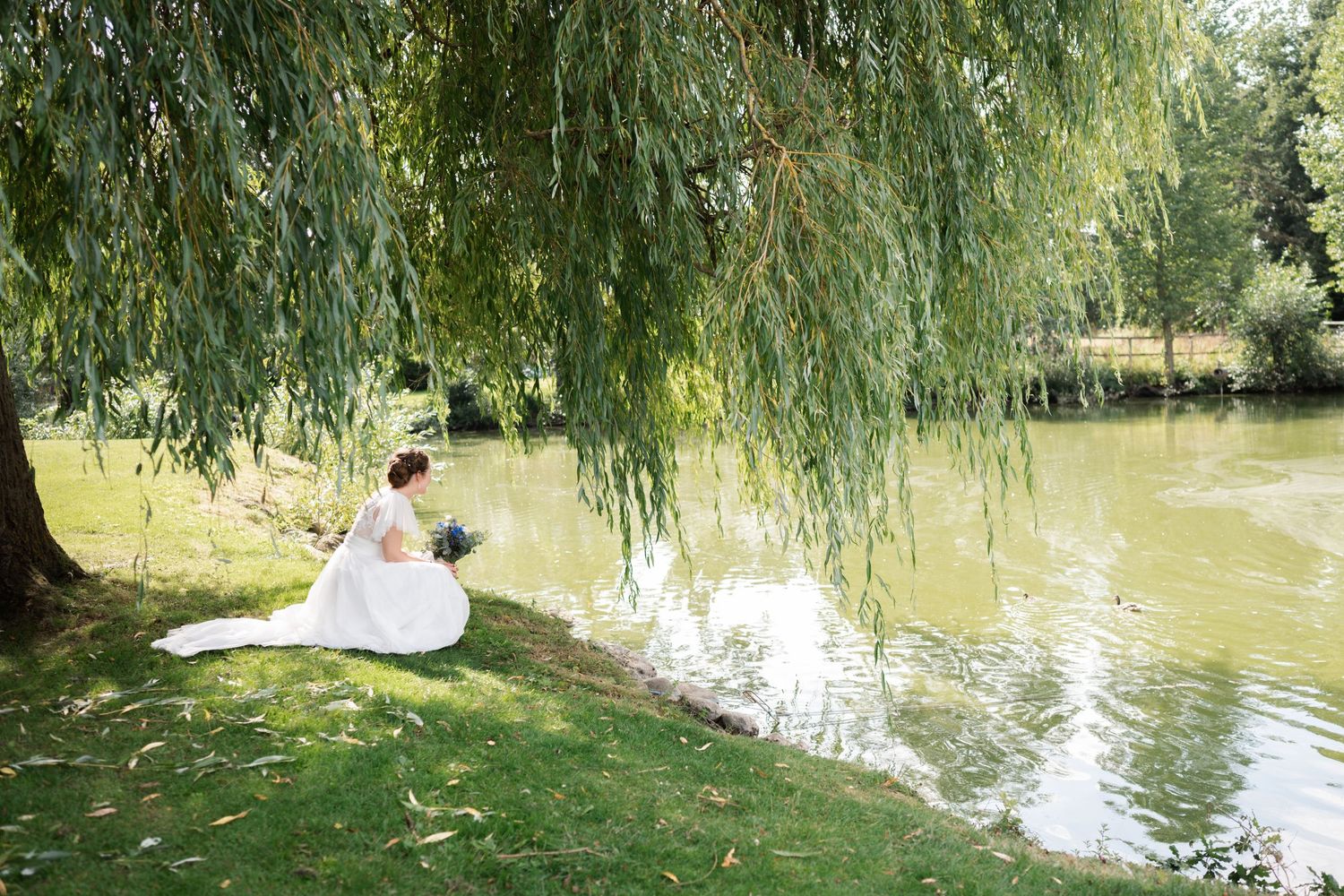Newlyweds sit under a weeping willow tree by a tranquil pond in a romantic outdoor setting.