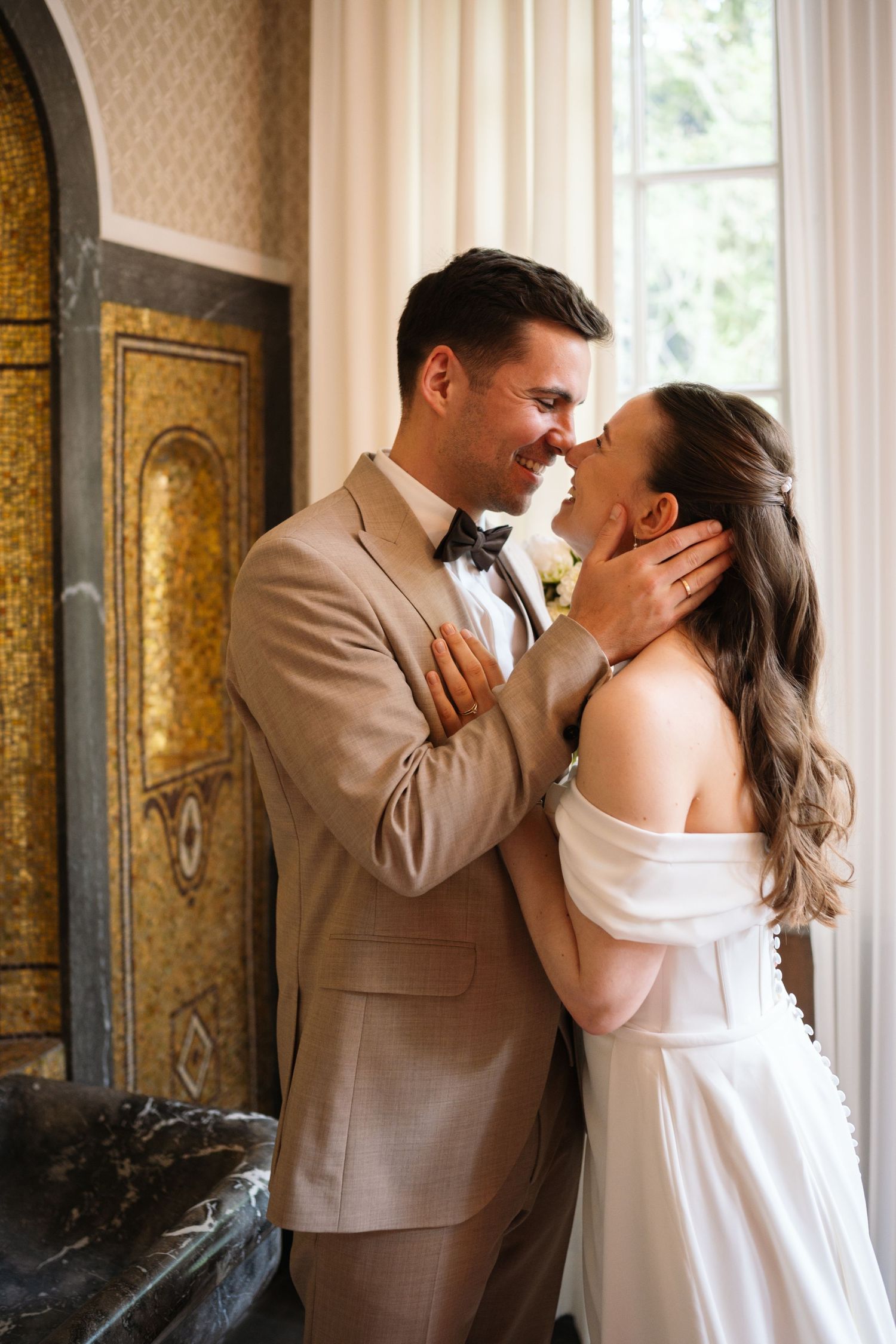A groom in a tan suit tenderly holds his bride's face as they share a romantic moment in an elegant venue.