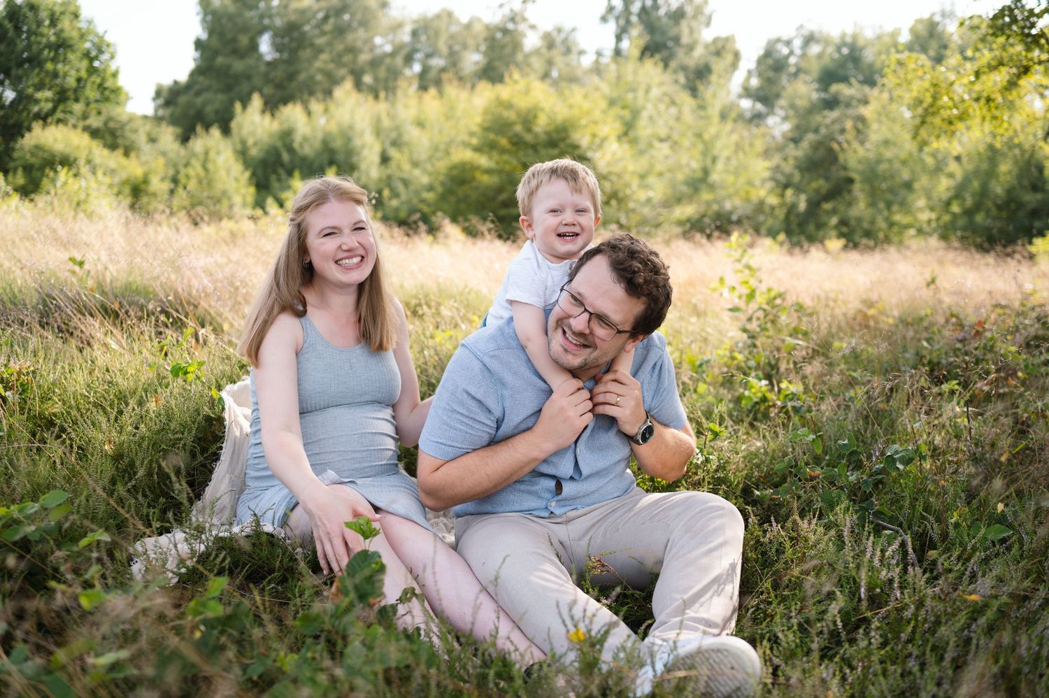 A couple shares joyful laughter while sitting together in a sunlit meadow with tall grass and wildflowers.