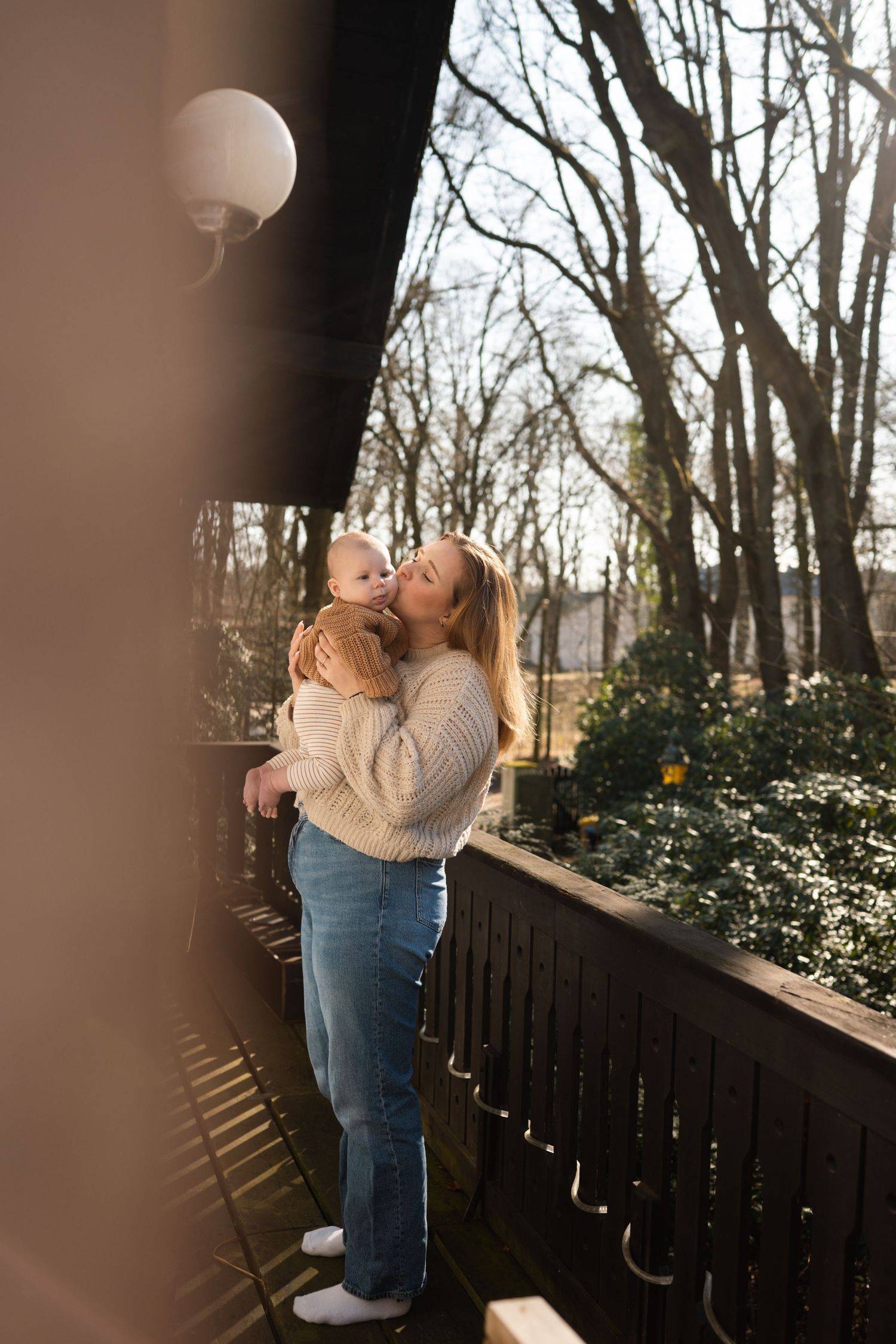 A mother holds her baby on a sunny balcony surrounded by bare trees and green shrubs in soft natural light.