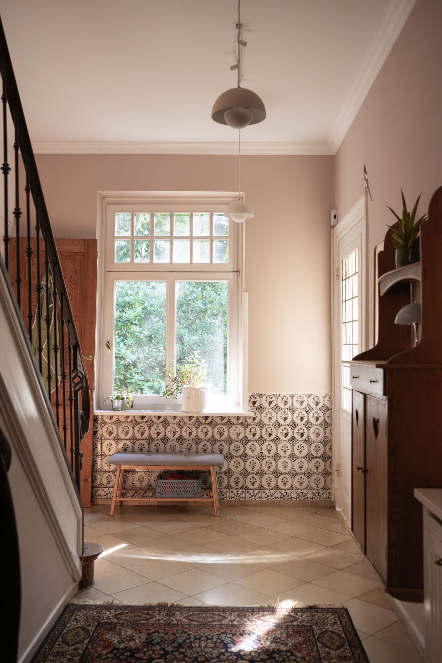 A bright entryway with pink walls features patterned tiles, wooden bench and natural light from a window.