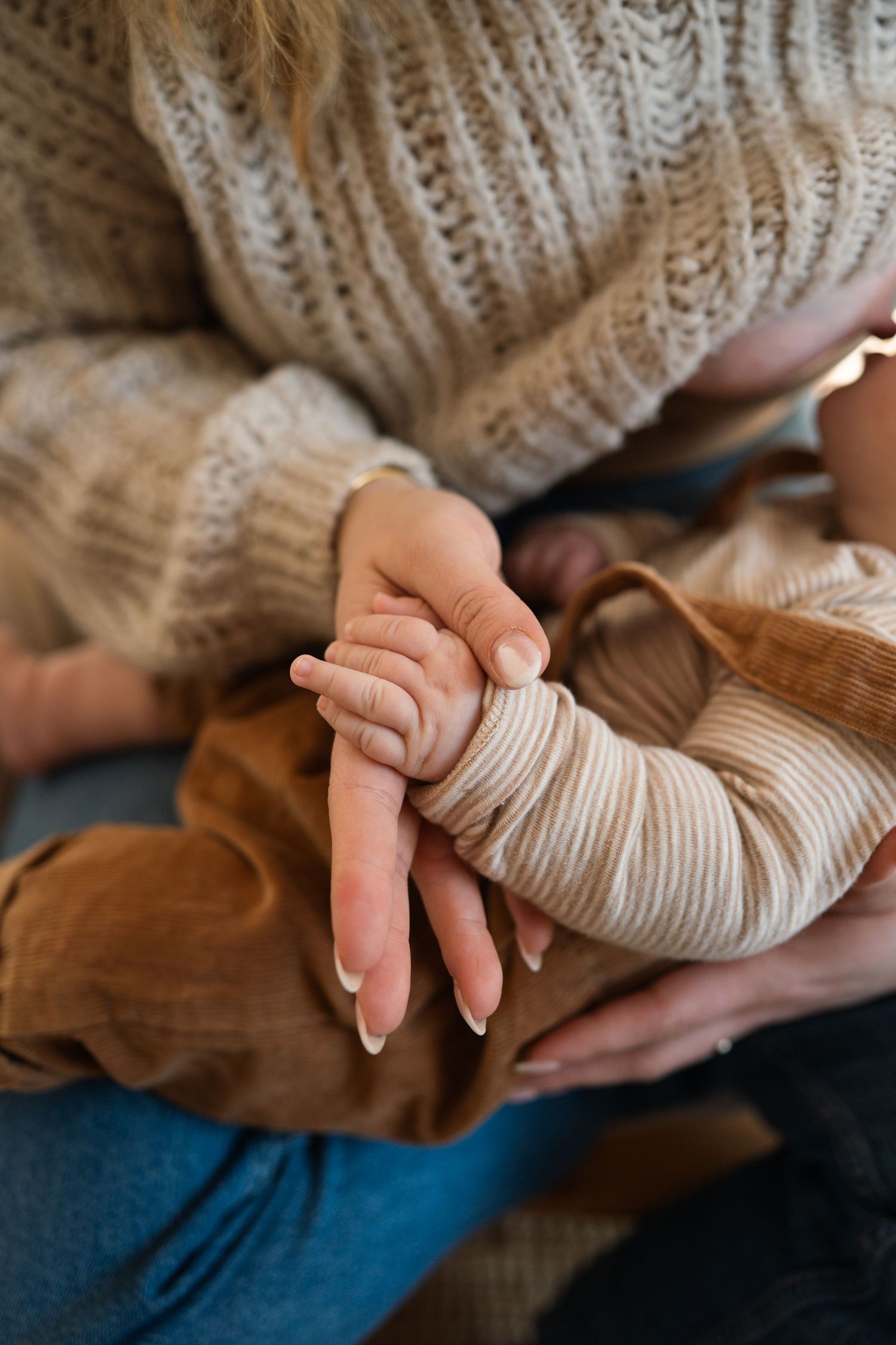 A mother in a cozy knit sweater gently holds her newborn baby's tiny hand, captured in a warm, intimate close-up.
