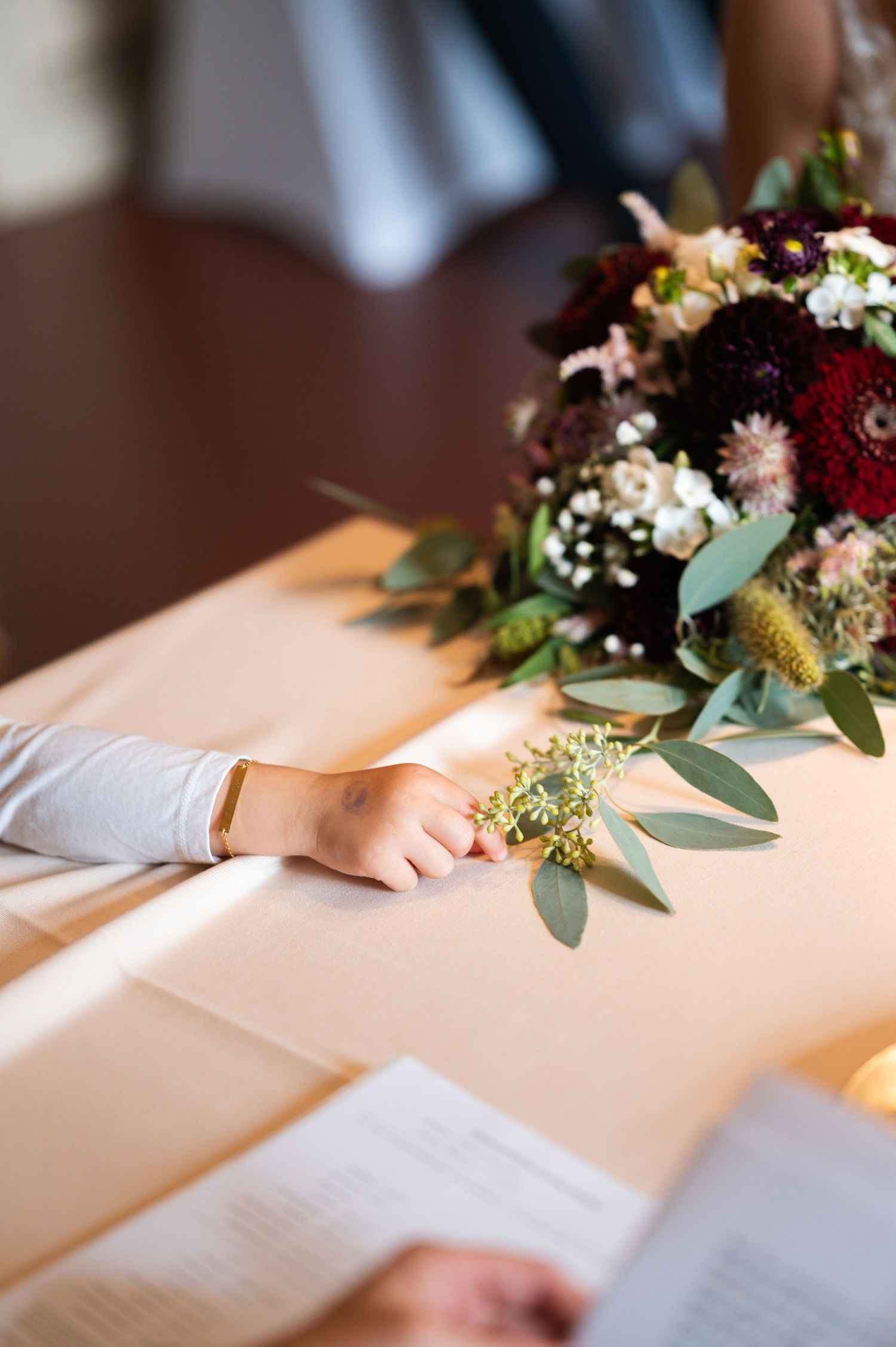 A beautiful burgundy and blush floral arrangement sits on a table next to hands holding papers during a ceremony.