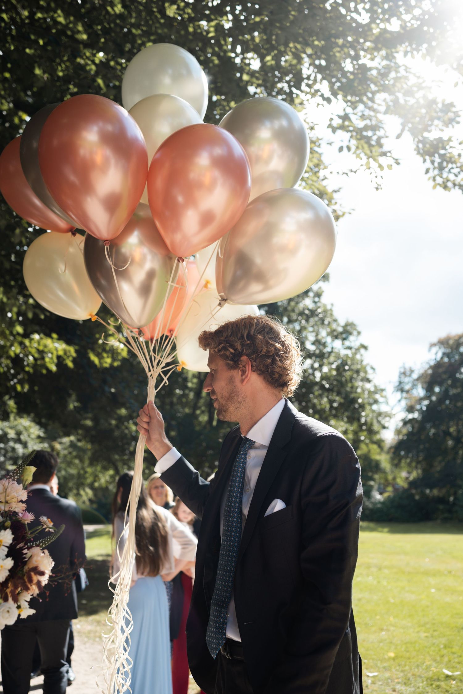 Wedding guests holding peach and white balloons in a sunny outdoor park setting.