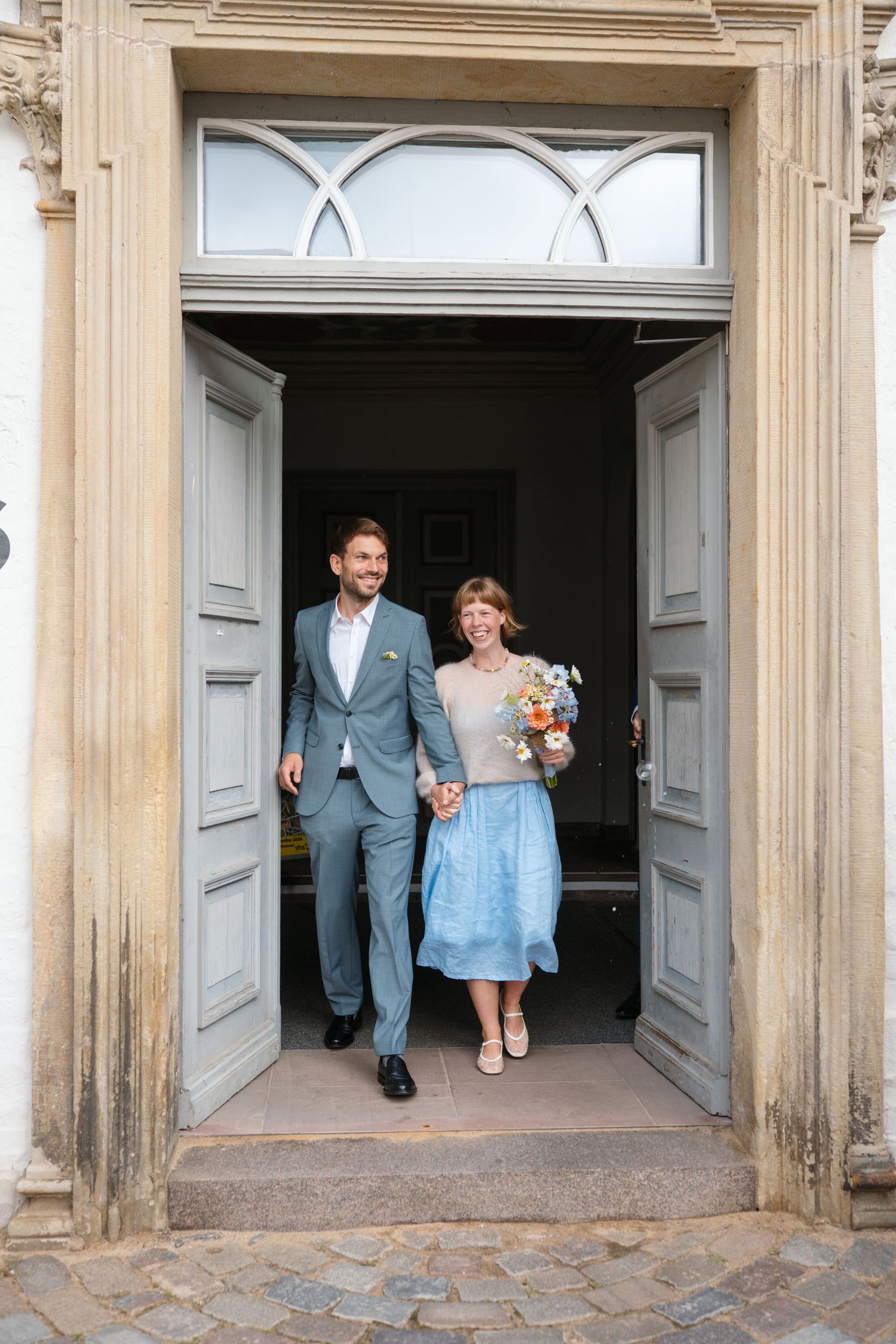 Couple exits elegant building doors, man in gray suit, woman in blue skirt holding colorful bouquet, celebrating wedding day.