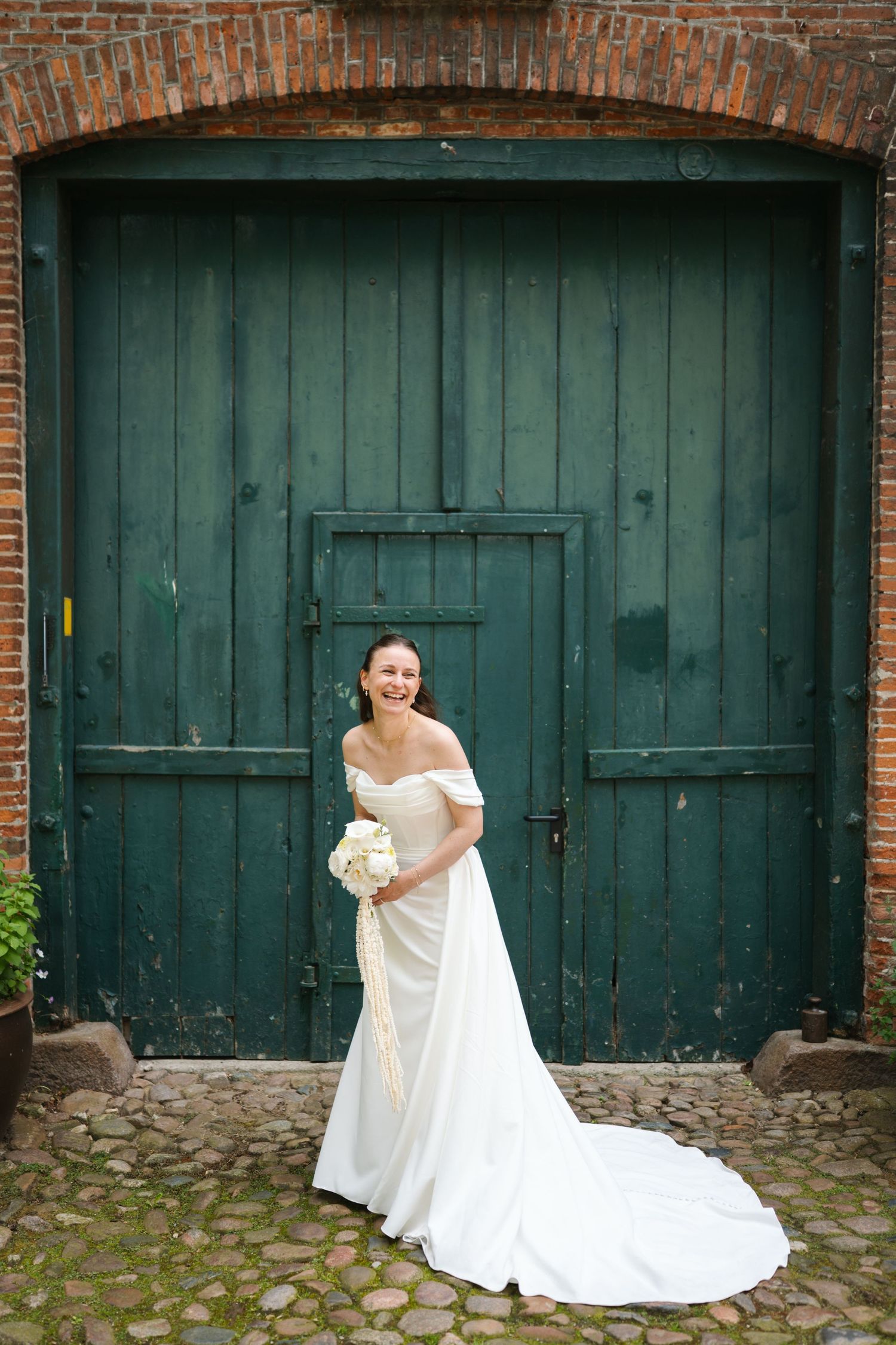 Elegant bride in white off-shoulder gown with train poses before rustic green wooden doors holding white bouquet.