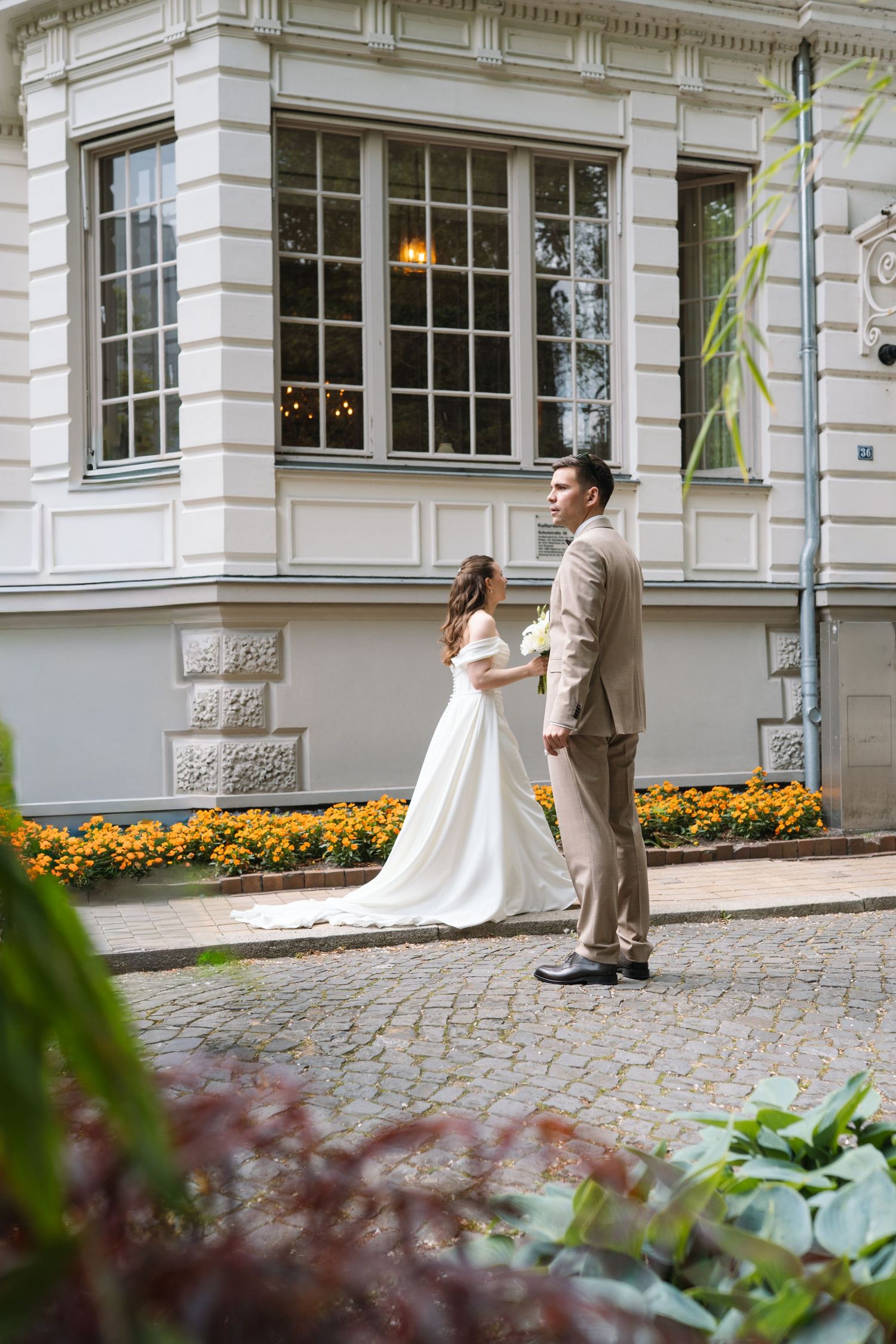 Bride and groom hold hands outside elegant building with orange flowers on cobblestone path.