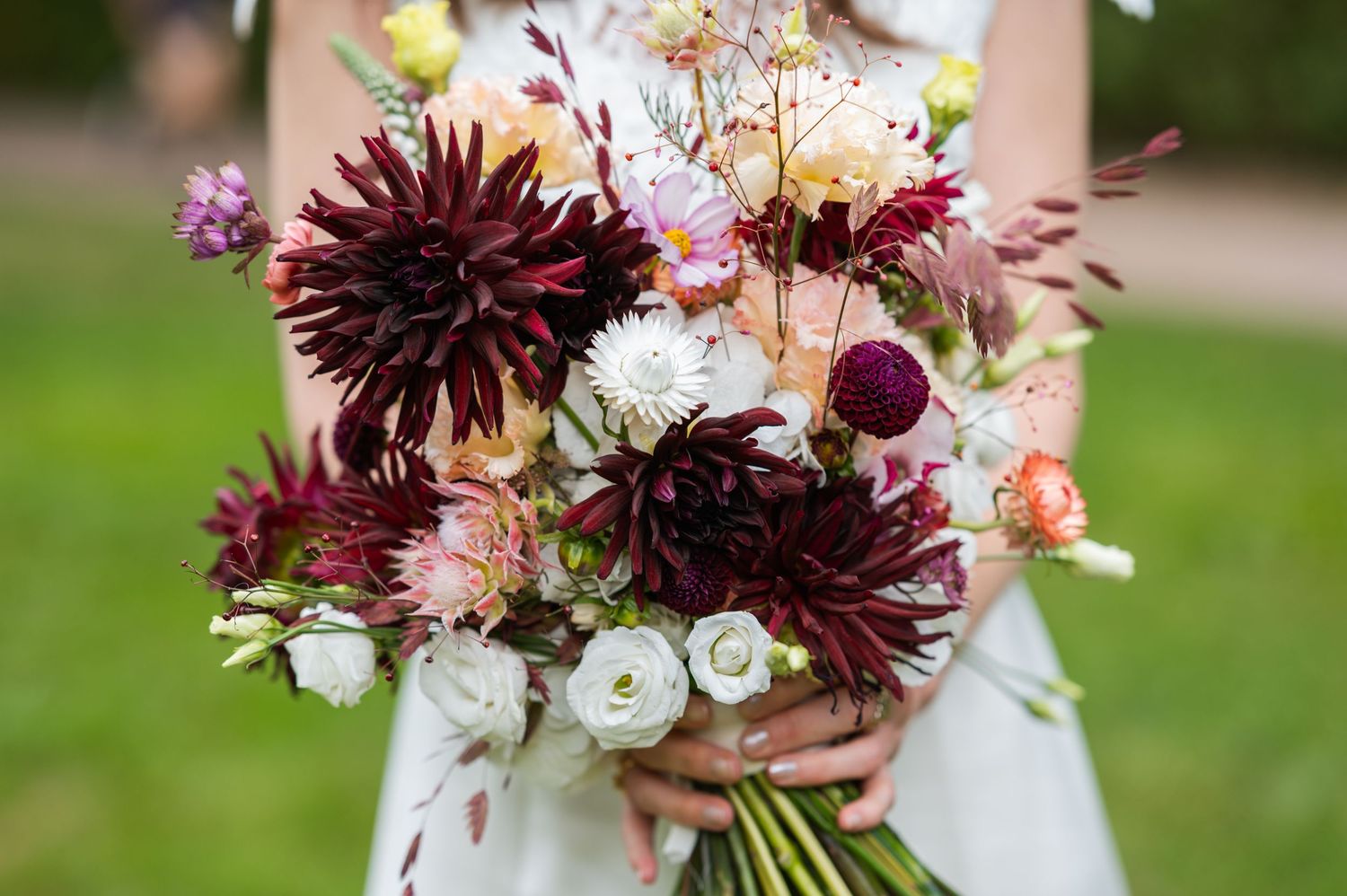 A romantic wedding bouquet featuring deep burgundy dahlias mixed with soft white and blush flowers against a blurred background.