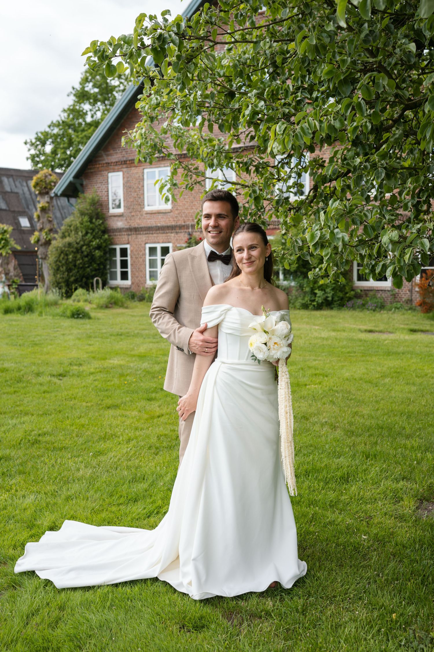 Bride in white off-shoulder gown holding bouquet stands with groom in tan suit on green lawn before brick building.
