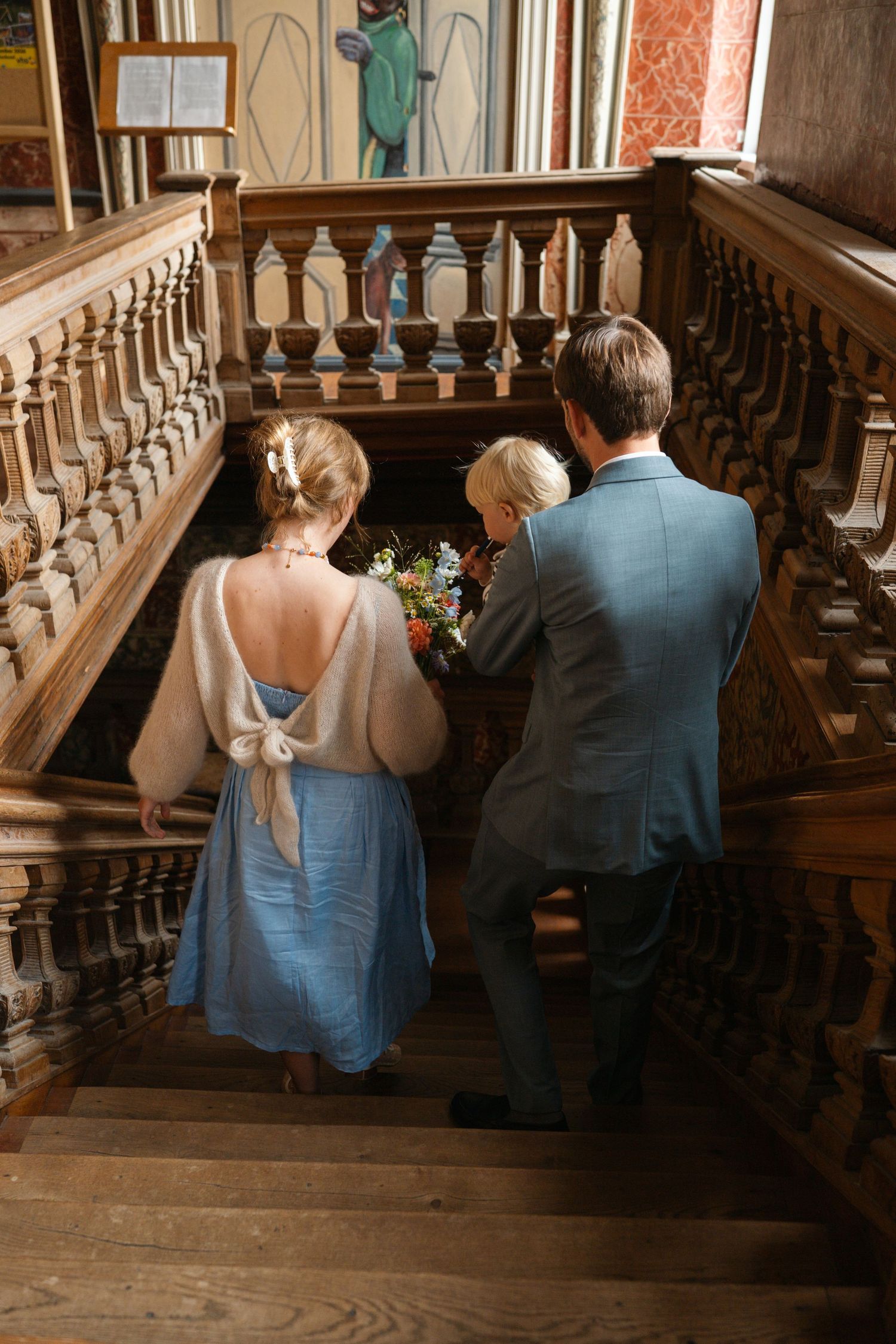 Bride and groom descending ornate wooden staircase, woman in blue dress with white shawl holding bouquet.