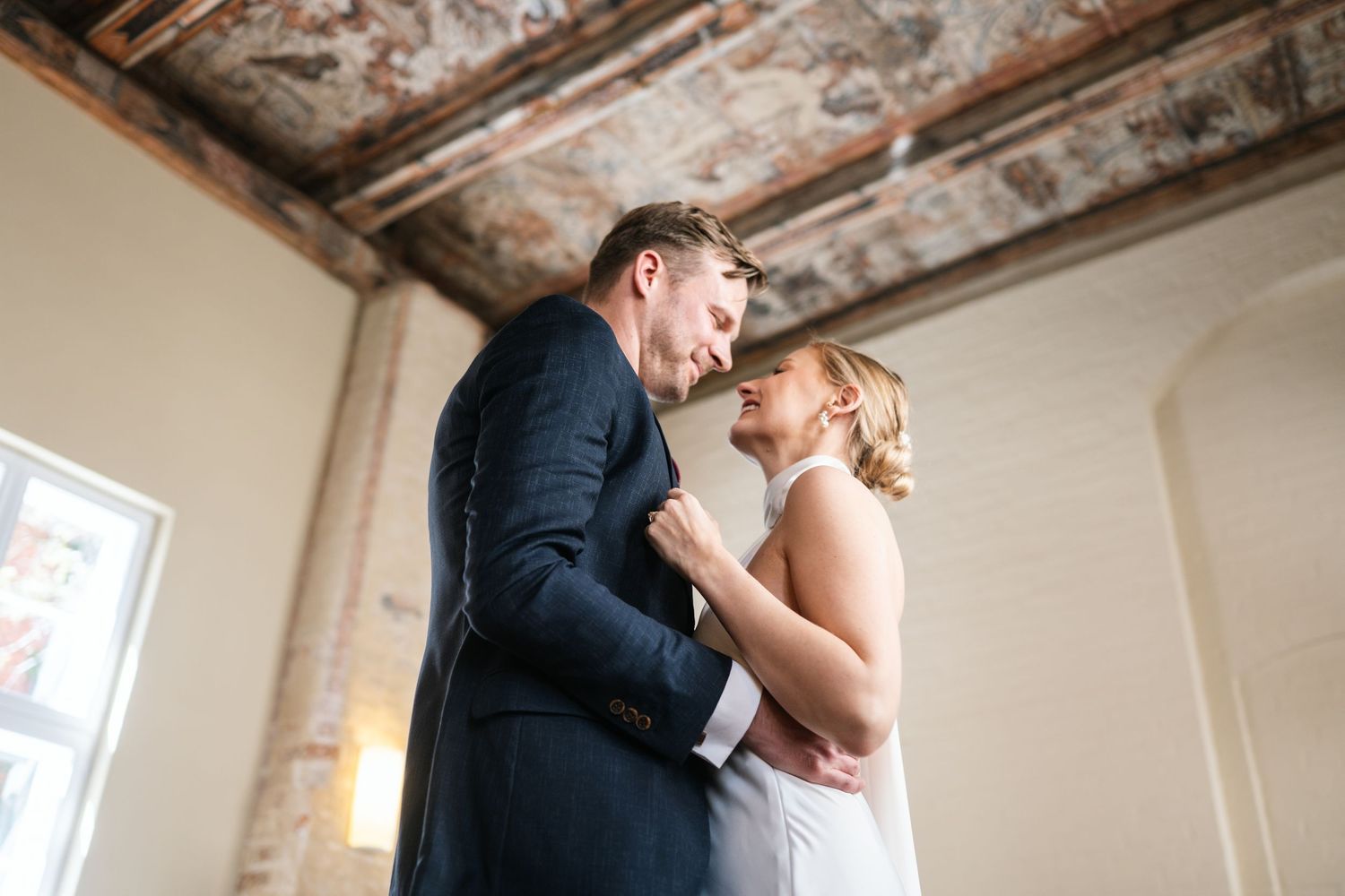 Bride and groom share a romantic moment in a rustic venue with exposed brick walls and ornate ceiling.