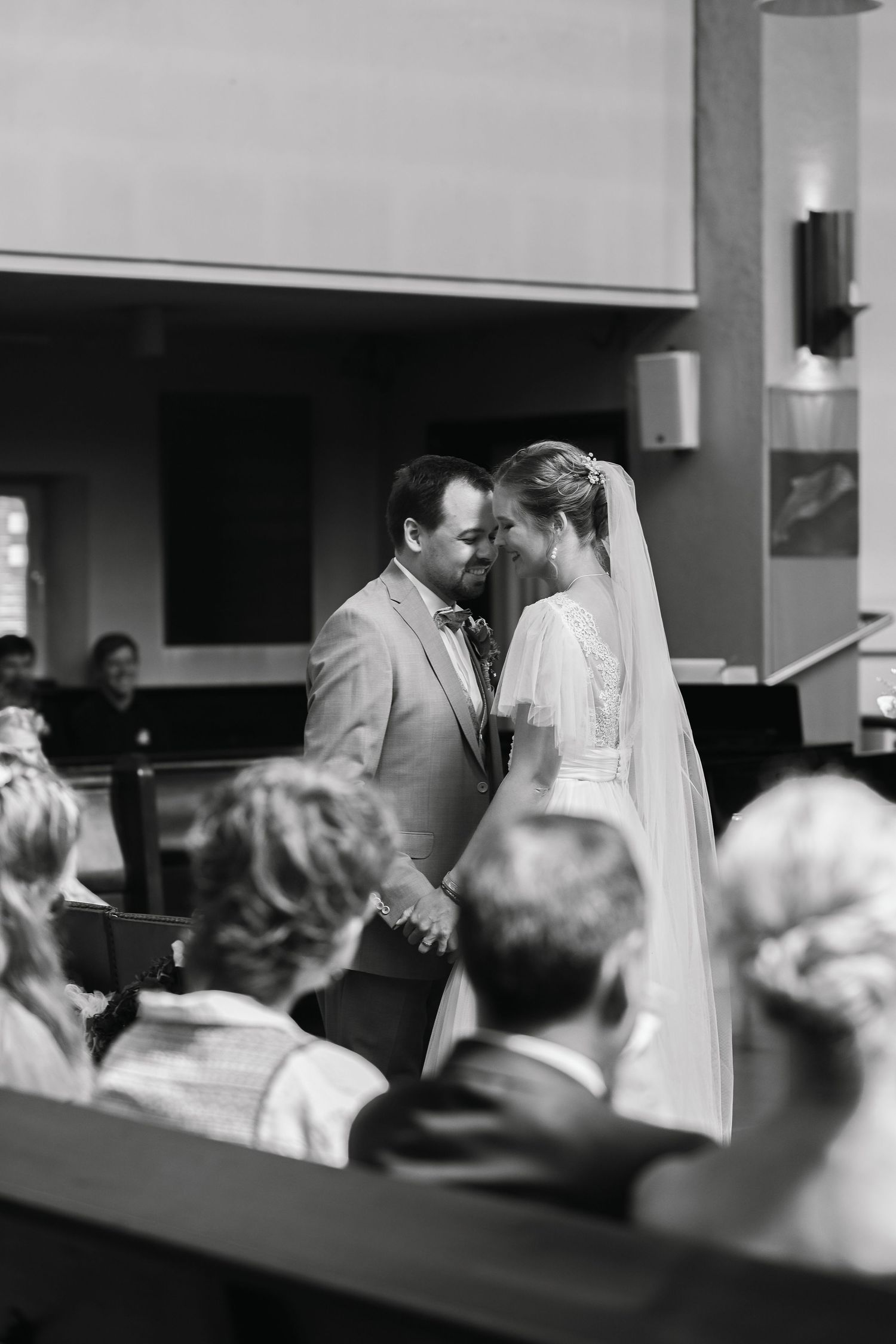 A black and white wedding ceremony photo showing a couple exchanging vows at the altar with guests in attendance.