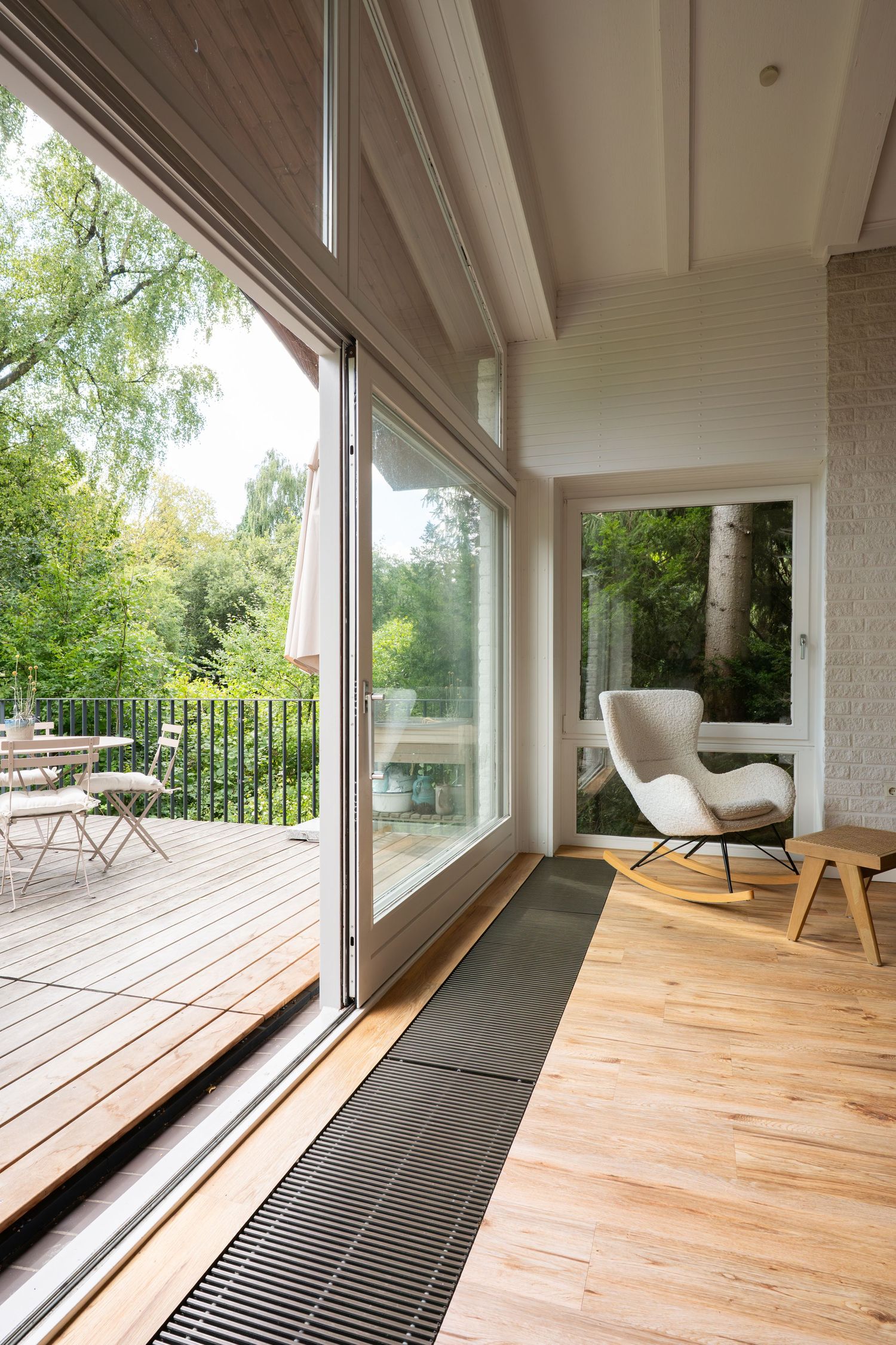 Modern living room with sliding glass doors opening to wooden deck, featuring a white lounge chair and floor heating vents.