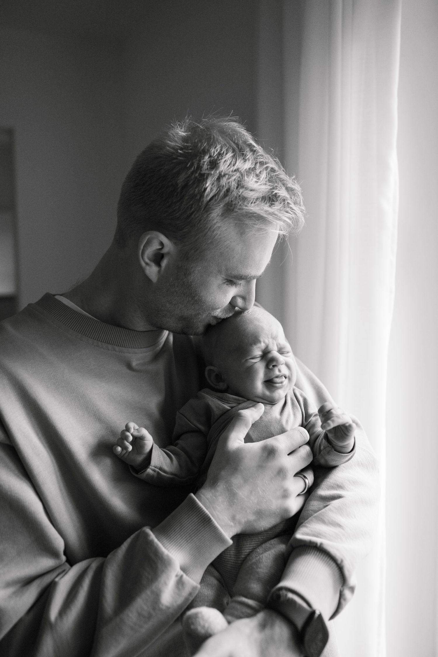 Black and white photo of a father tenderly kissing his newborn baby held close to his chest near a window.