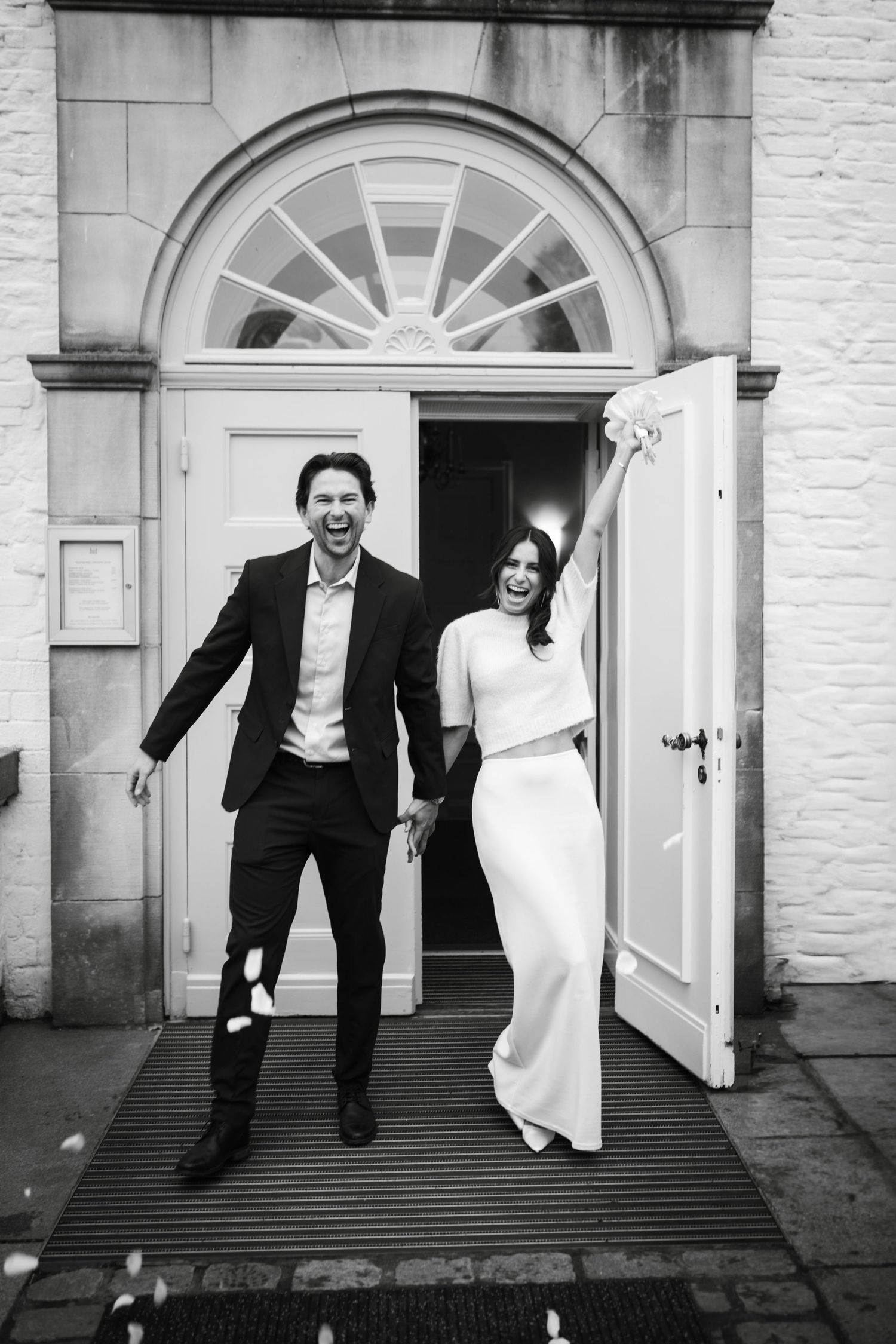 A joyful couple in formal attire runs and leaps together outside an elegant building entrance with white doors.