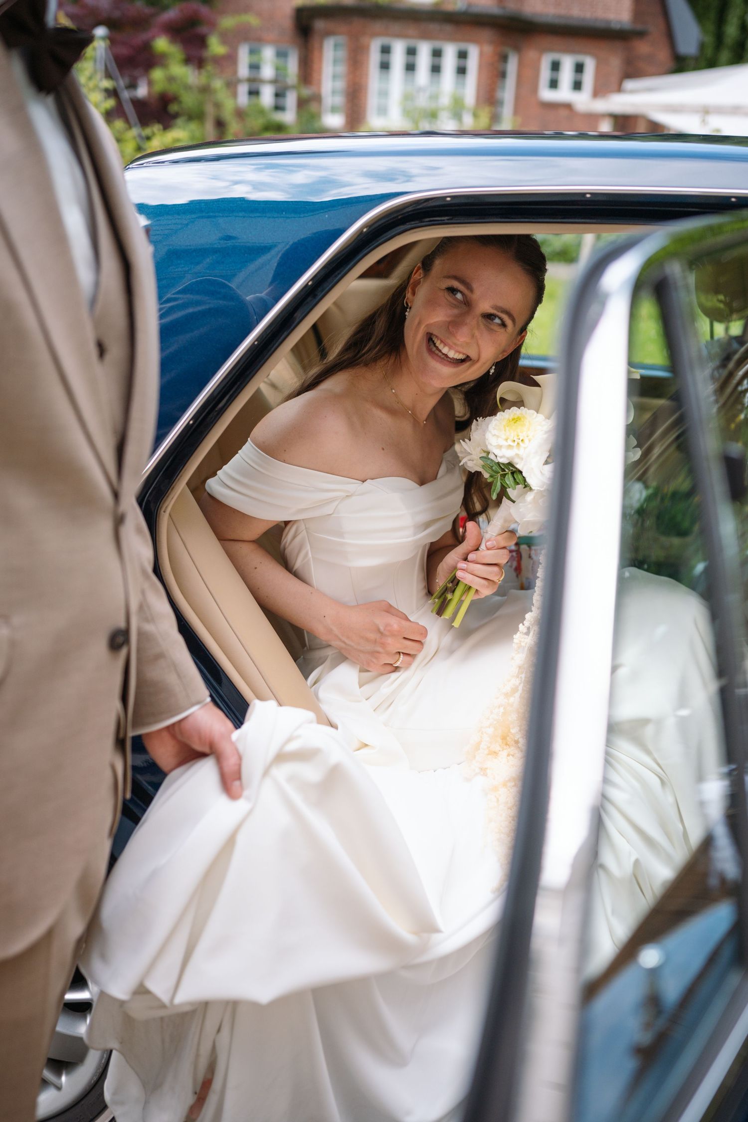 Smiling bride in off-shoulder white gown holding white bouquet stepping out of vintage blue car on wedding day.